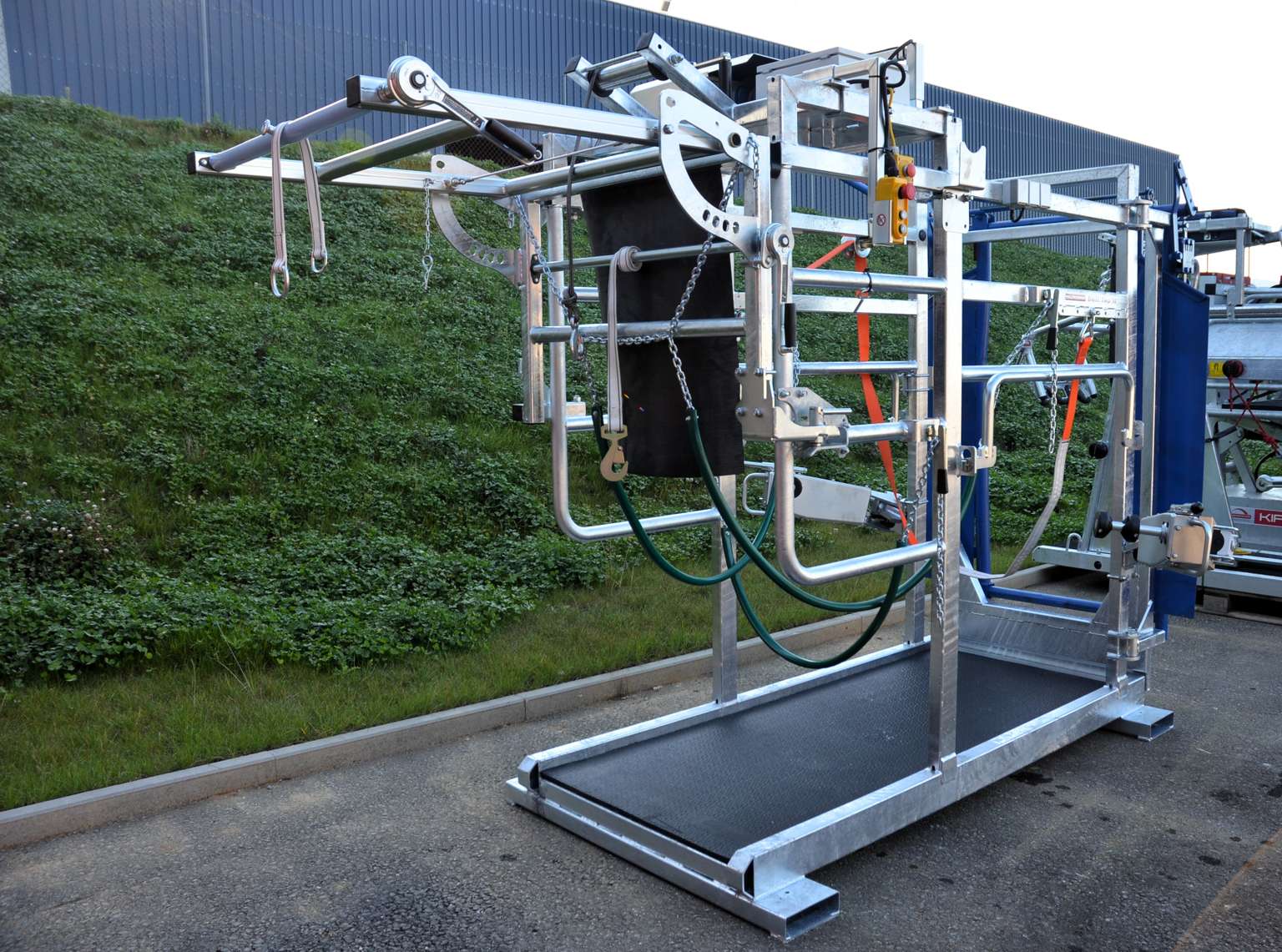 Metal livestock handling equipment on a paved surface, featuring various mechanical components and a green hill in the background.