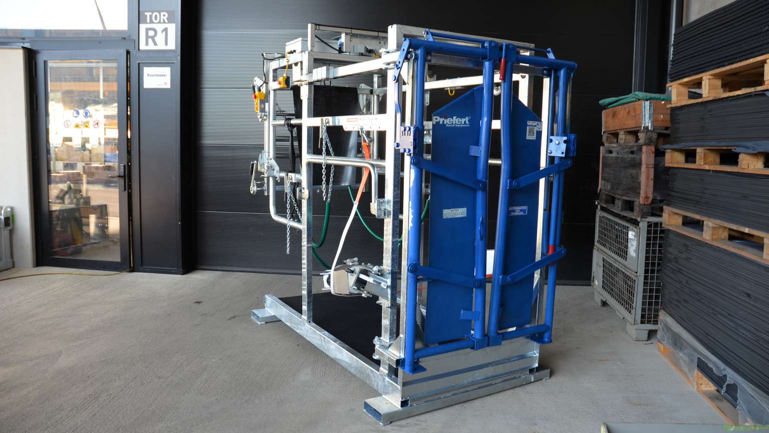 A blue industrial machine with metal framework and cables stands on a concrete floor inside a warehouse.