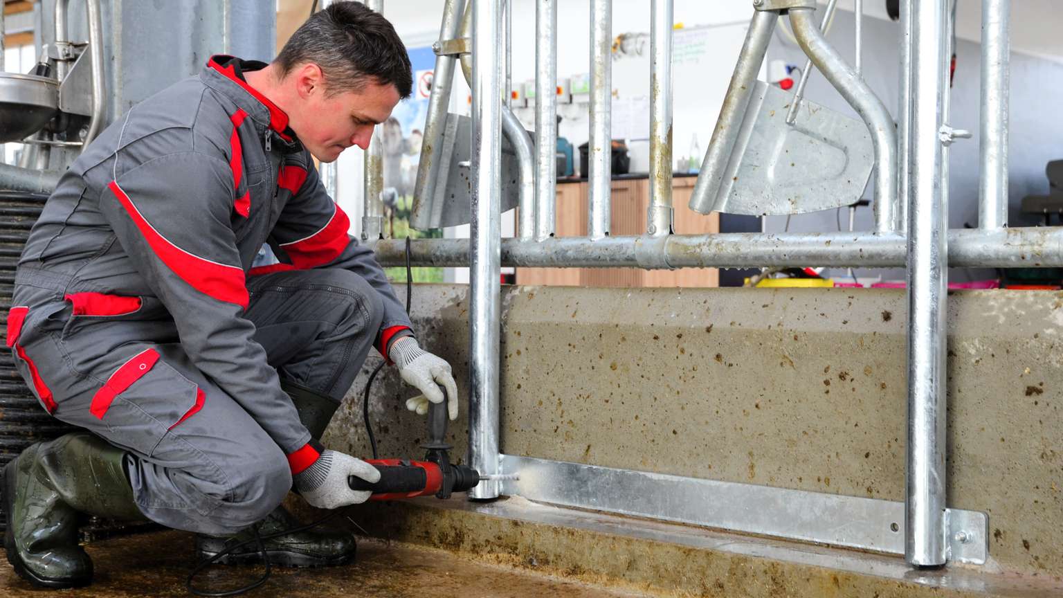 Worker in gray and red overalls uses a power tool to adjust metal bars on a concrete platform in an industrial setting.