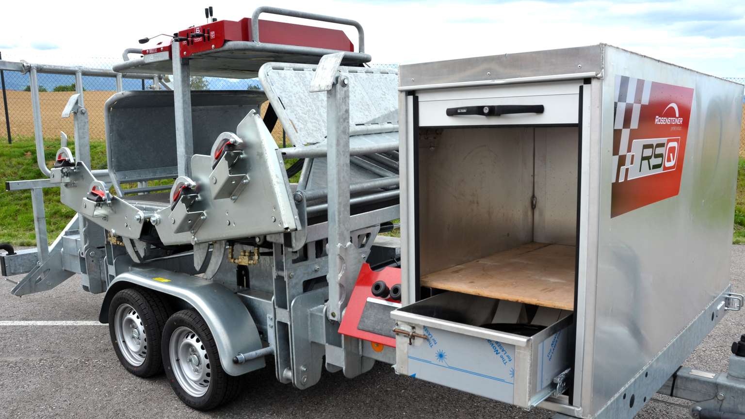 A transport trailer with equipment, featuring an open storage compartment and mounted machinery, parked on a paved surface.