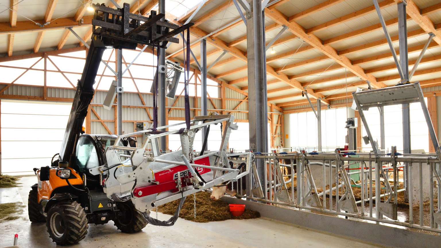 A tractor with a front-mounted machine operates inside a modern barn, moving materials into feeding troughs for livestock.