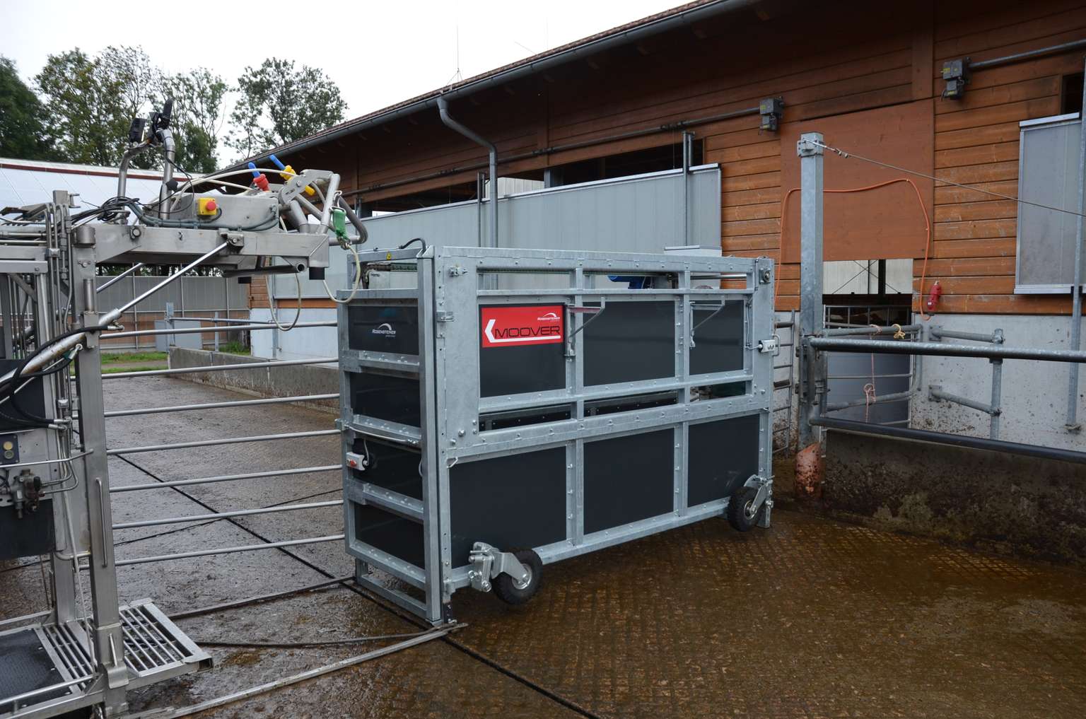 A metal livestock handling crate on wheels is positioned near a barn building, with various equipment connected to it.