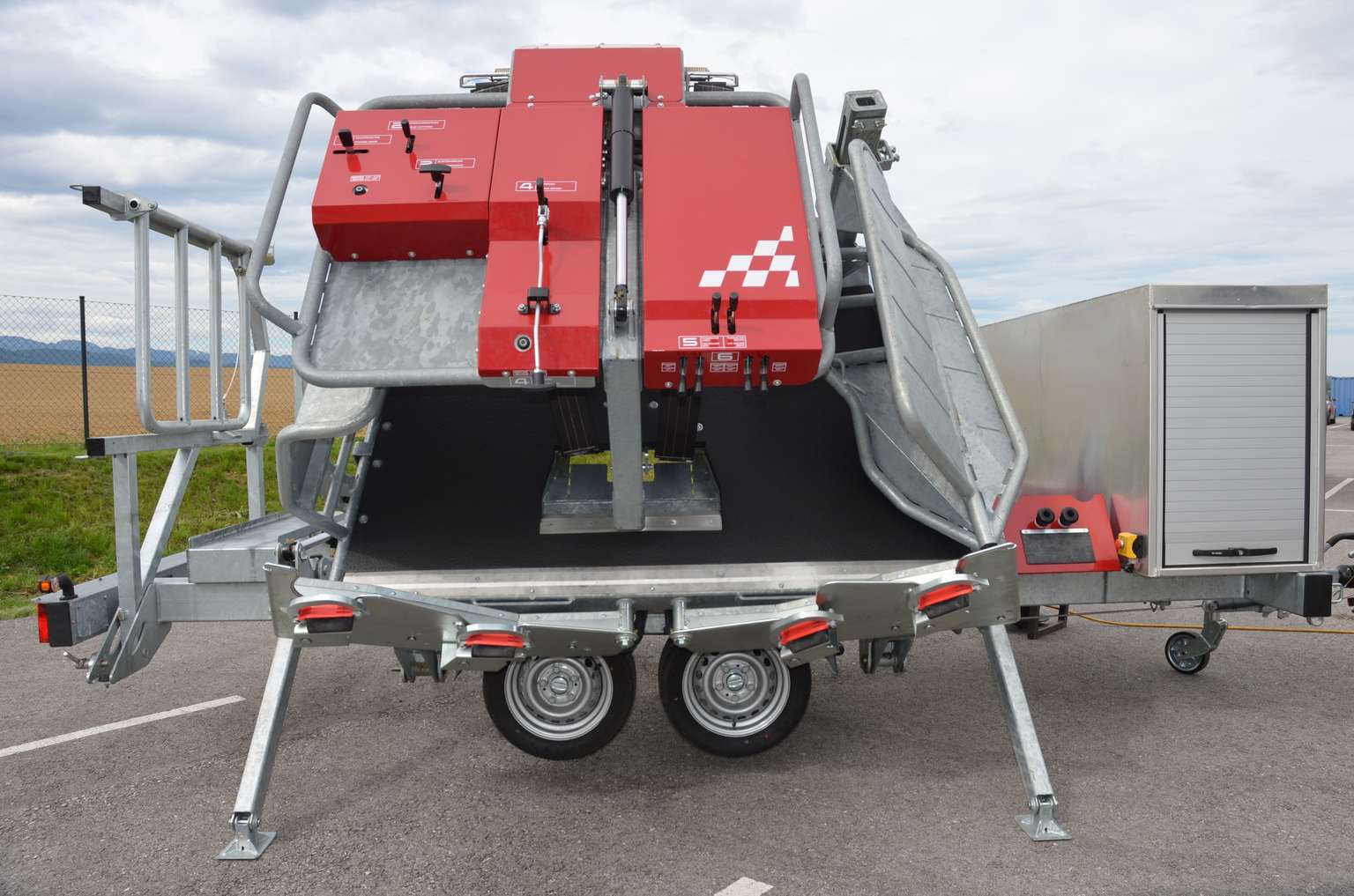 Red and silver livestock handling trailer with metal bars and wheels, parked on a paved area outdoors.