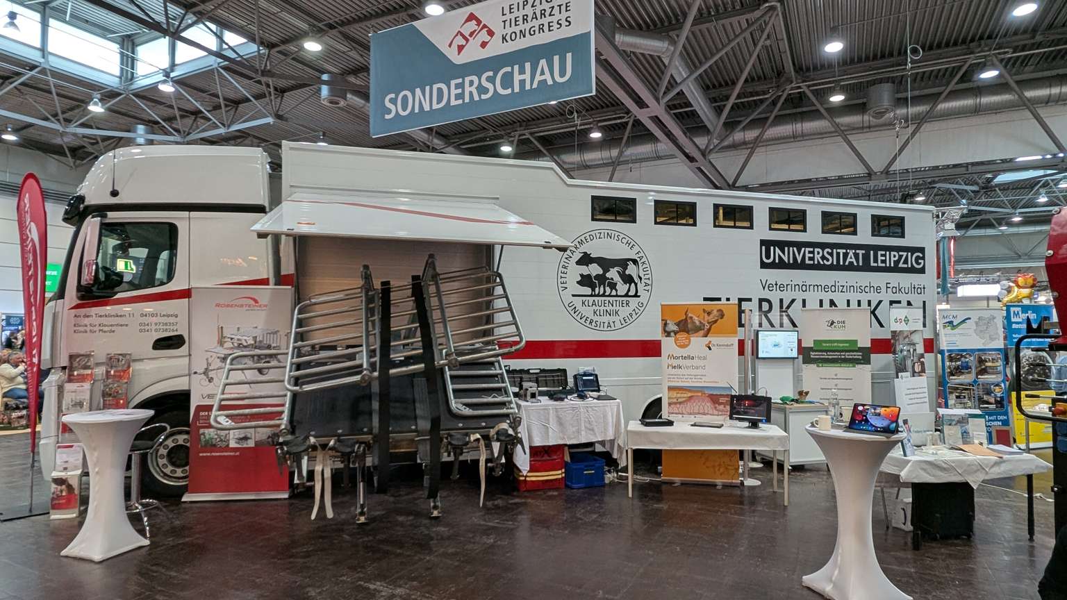 Exhibition setup with a university truck, display boards, and informational materials inside a large indoor hall, under a sign reading "Sonderschau."