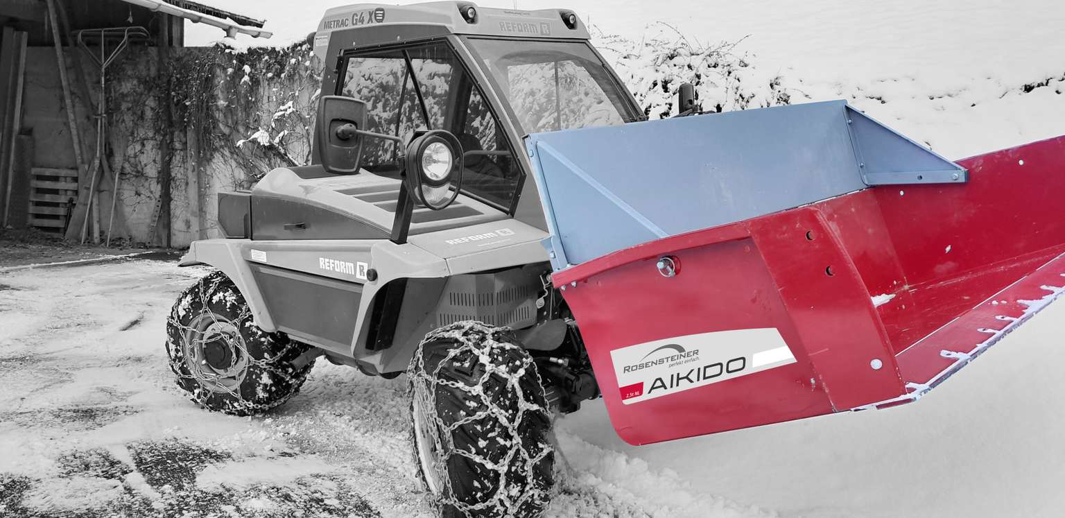 A tractor equipped with a red and blue AIKIDO snow plow attachment clears snow from a path, surrounded by a snowy landscape.