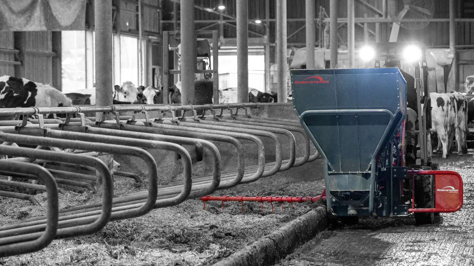 Feeding machine in a barn, distributing feed to cows in stalls. Dimly lit with some colored accents on the machine.