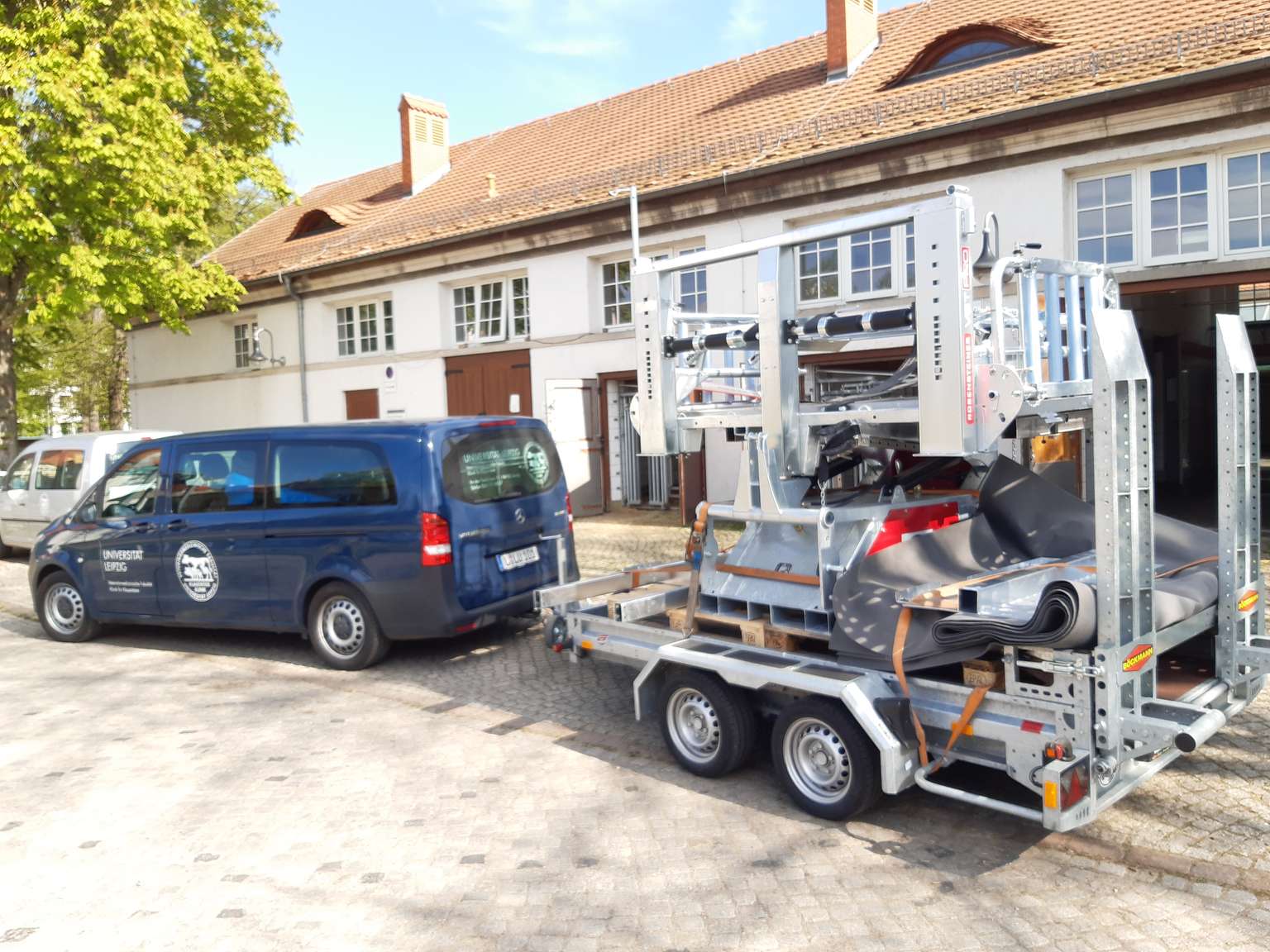 A blue van is parked outside a building, towing a trailer loaded with machinery, under a clear sky.
