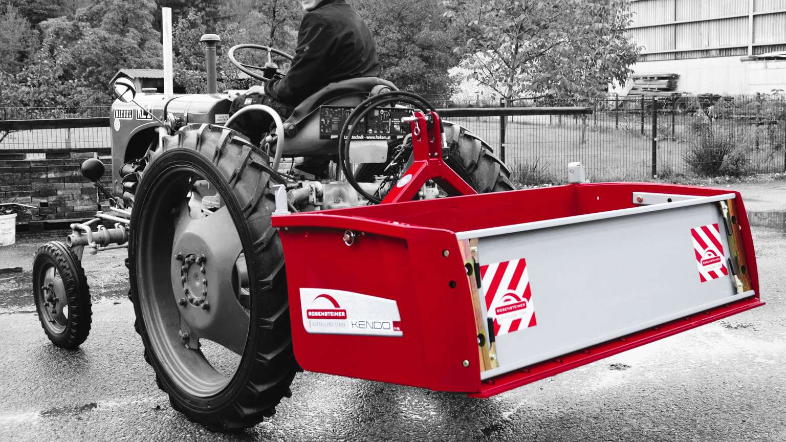 A person on a tractor with a large red attachment on the back, set against a grayscale background with trees and a building.