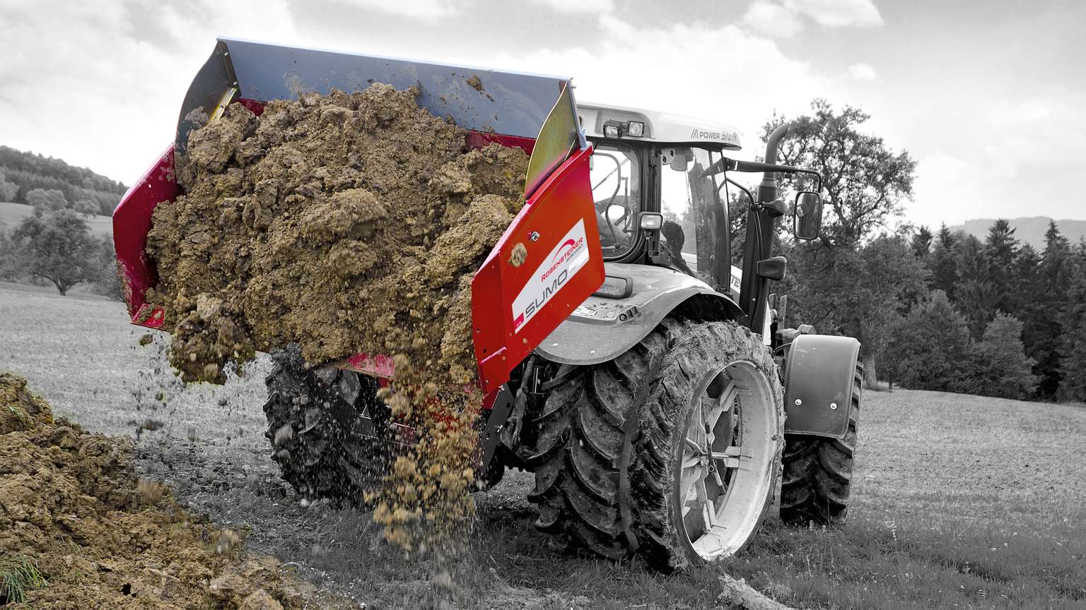 A tractor is spreading manure on a grassy field, with a red attachment on the back. Trees and hills are visible in the background.
