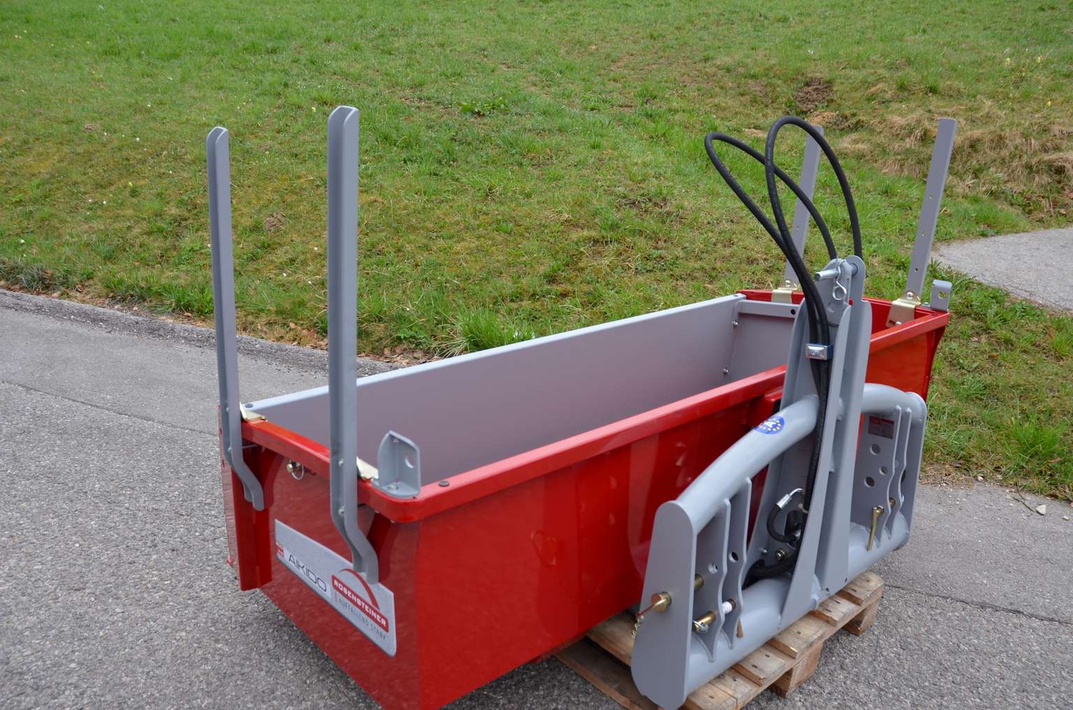 Red metal attachment for a tractor with arm-like structures and black cables, placed on a wooden pallet near a grassy area and pavement.