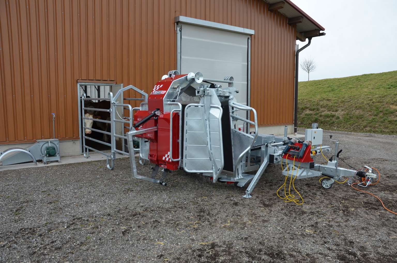 Red and silver cattle grooming machine outside a barn, featuring metal framework and various cables, set on a gravel surface with a grassy hill nearby.