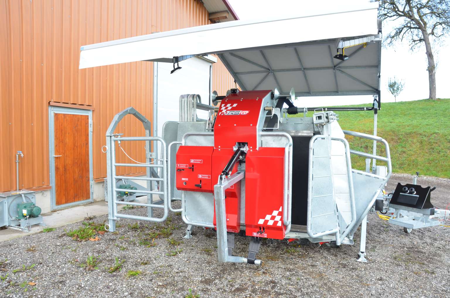 Red and silver automated milking machine outside a barn with a gated enclosure and a metal canopy.