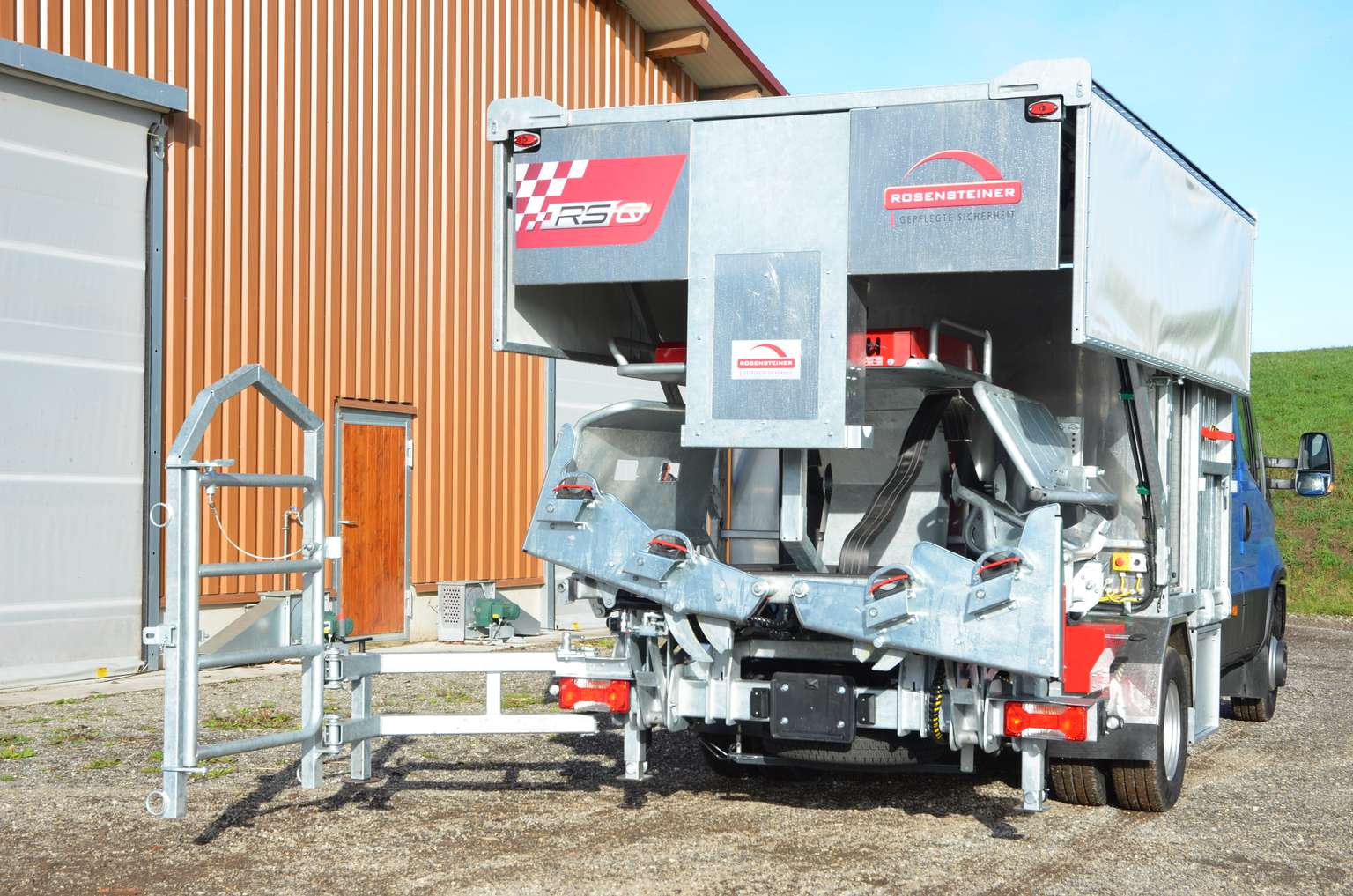 Large agricultural machine parked on concrete, with metal components, next to a red building. Blue sky and grass in the background.