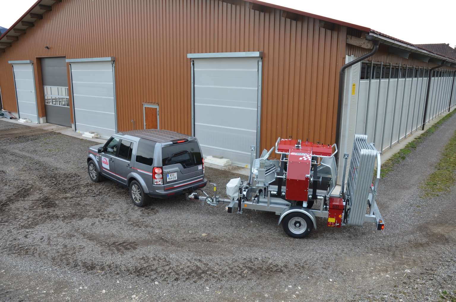 SUV towing a portable wood processor on a gravel path beside a large barn with multiple closed garage doors.
