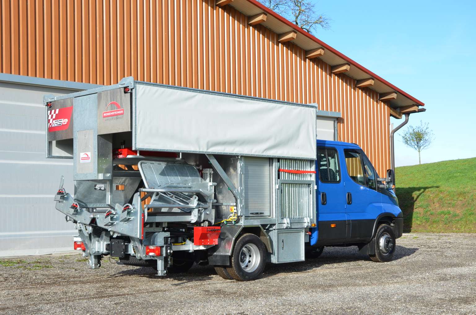 Blue and silver utility vehicle with attached machinery parked in front of a corrugated metal building on a clear day.