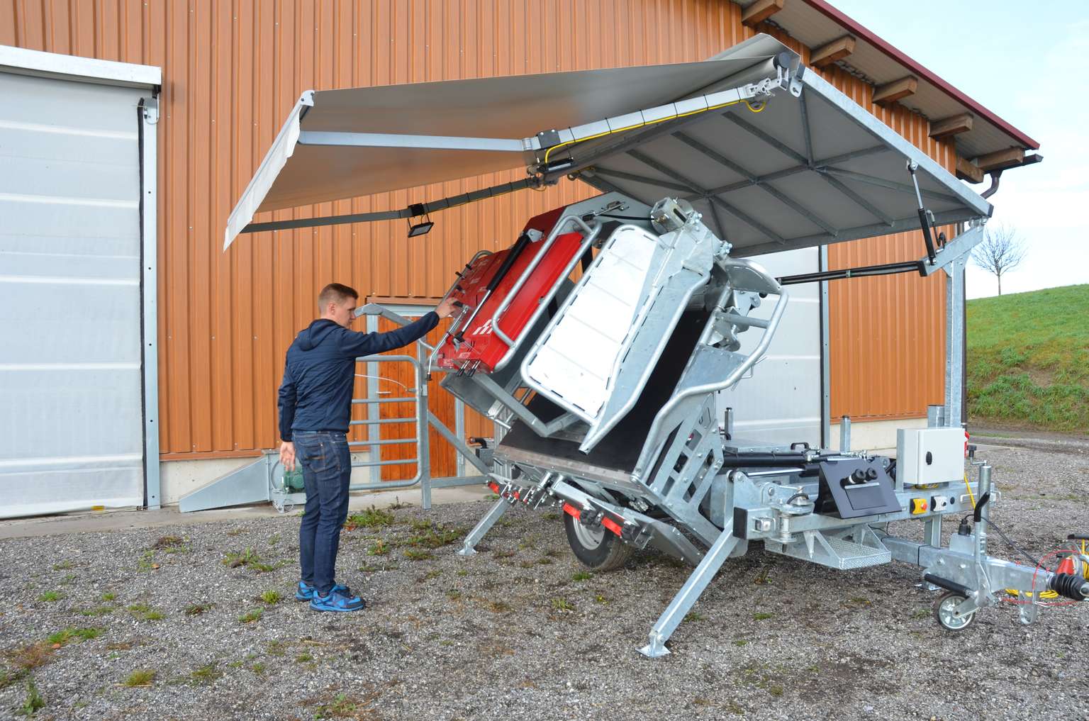 A person operates machinery beneath a metal canopy, next to a tilted, red agricultural machine beside a building.