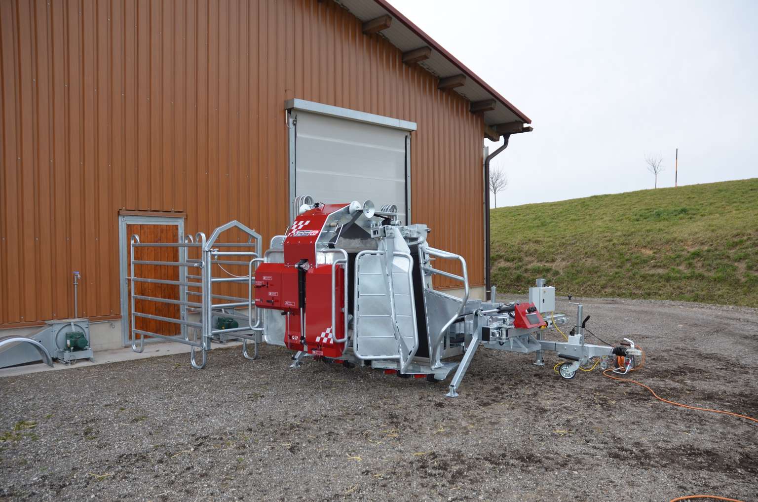 Red and silver farm machinery parked on gravel beside a beige building with a closed garage door.