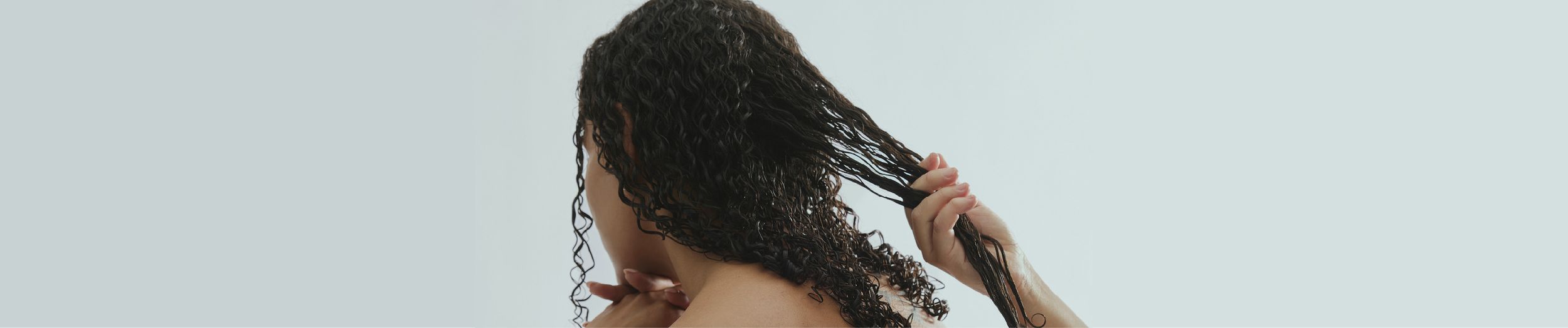 Person with wet, curly hair seen from behind, gently separating strands with hands against a light gray background.