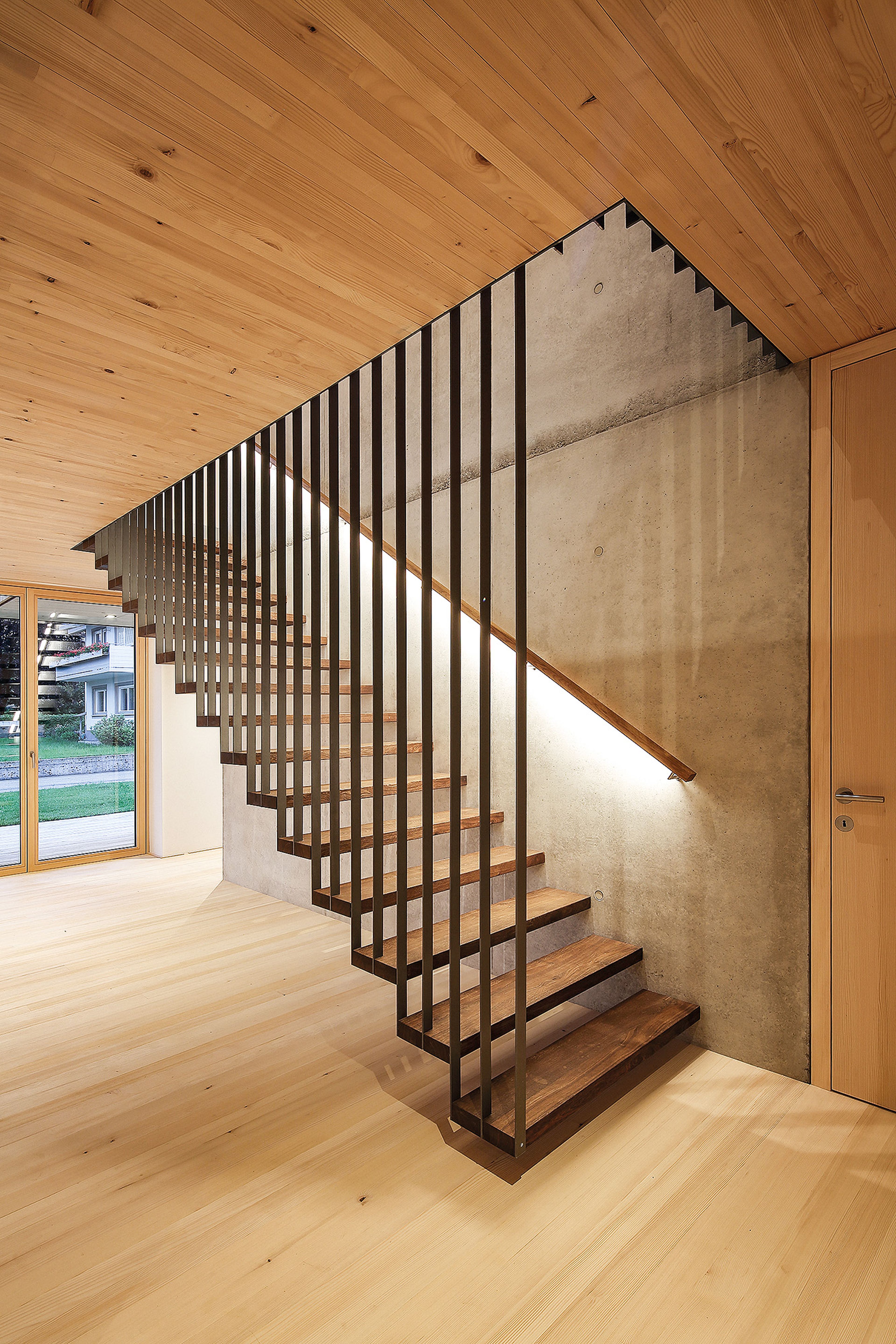 Moderne Holztreppe mit Metallgeländer vor einer Betonwand in einem minimalistischen Interieur mit großen Fenstern und hellem Holzboden.