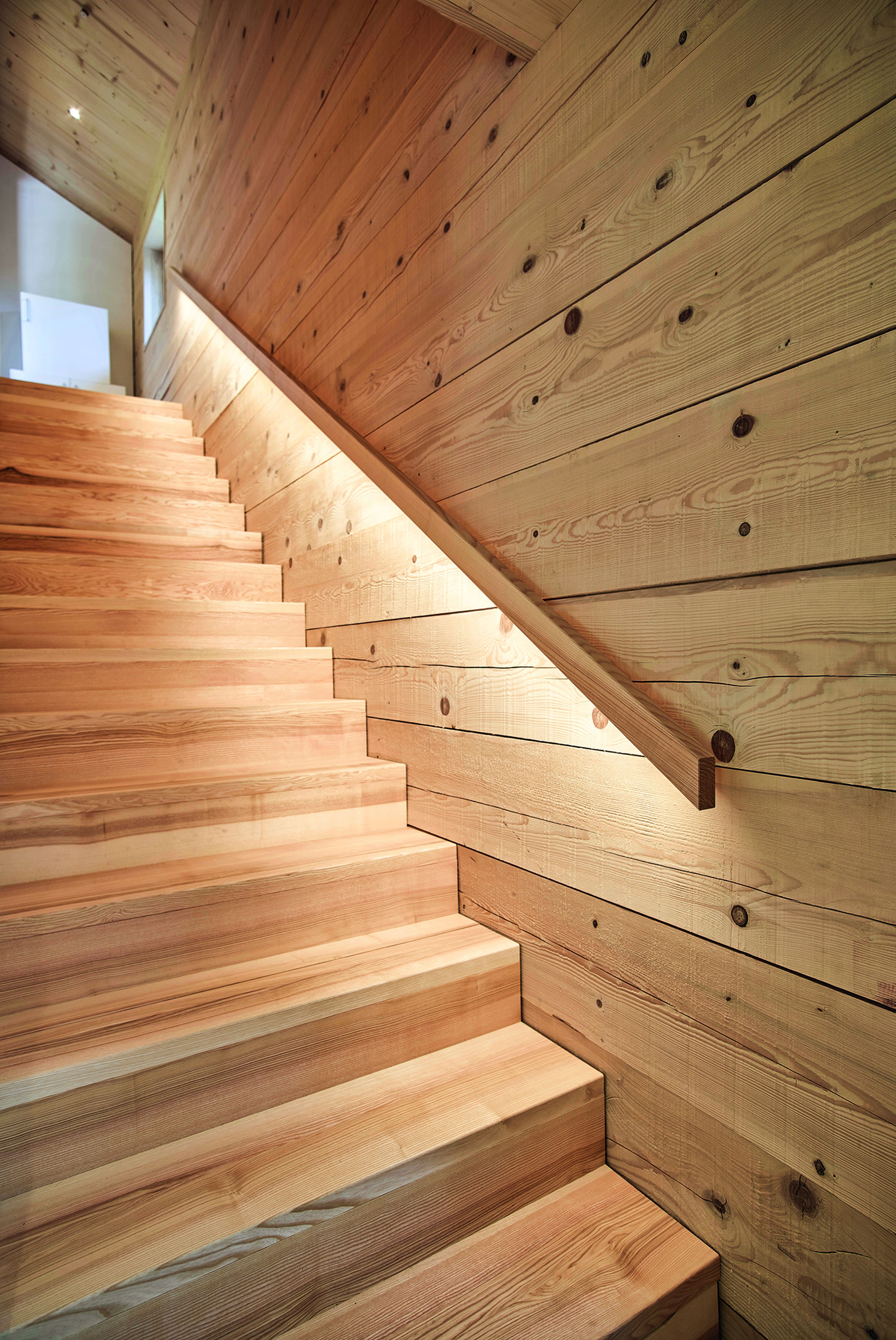Eine Holztreppe mit nahtlosen Seitenwänden aus horizontalen Holzpaneelen, die von unten sanft beleuchtet werden und so eine warme, einladende Atmosphäre schaffen.