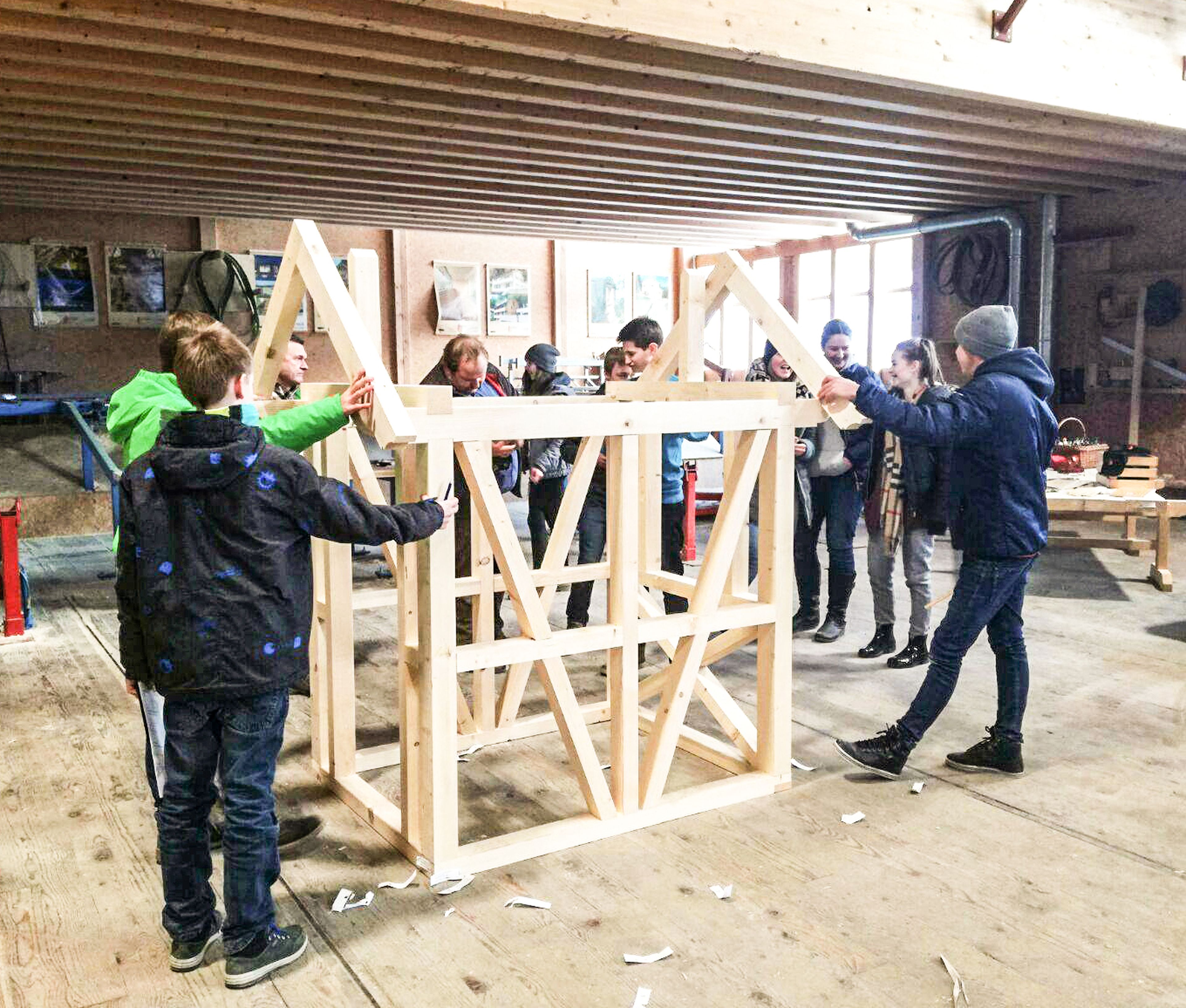 In einer geräumigen Werkstatt arbeitet eine Gruppe von Schulkindern gemeinsam am Aufbau einer Holzkonstruktion. Holzspäne liegen verstreut auf dem Boden.