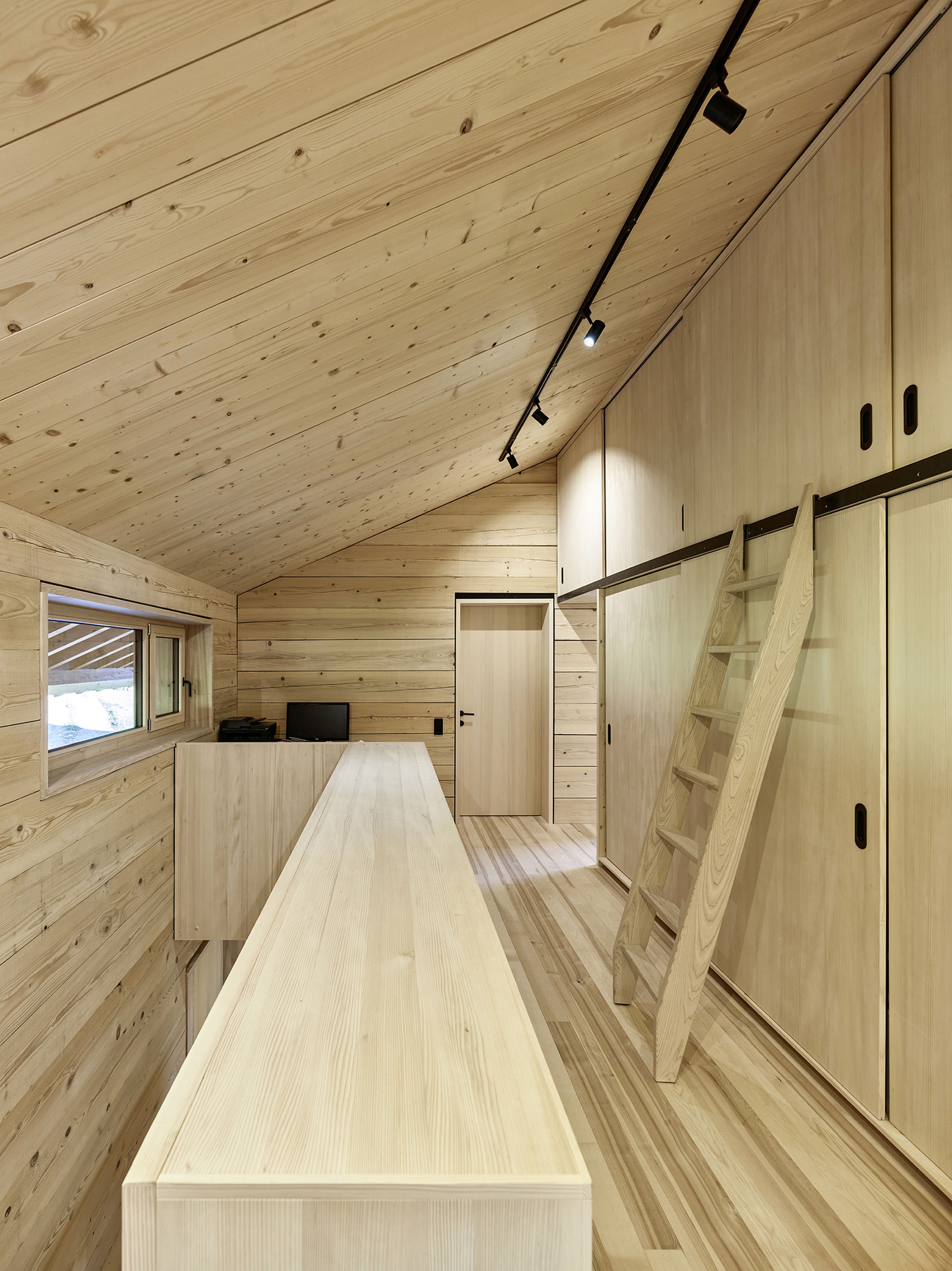 Gemütliches Dachzimmer mit Holzvertäfelung, Dachschräge, Einbauschränken, Leiterzugang und Schienenbeleuchtung. Durch ein Seitenfenster strömt natürliches Licht herein.