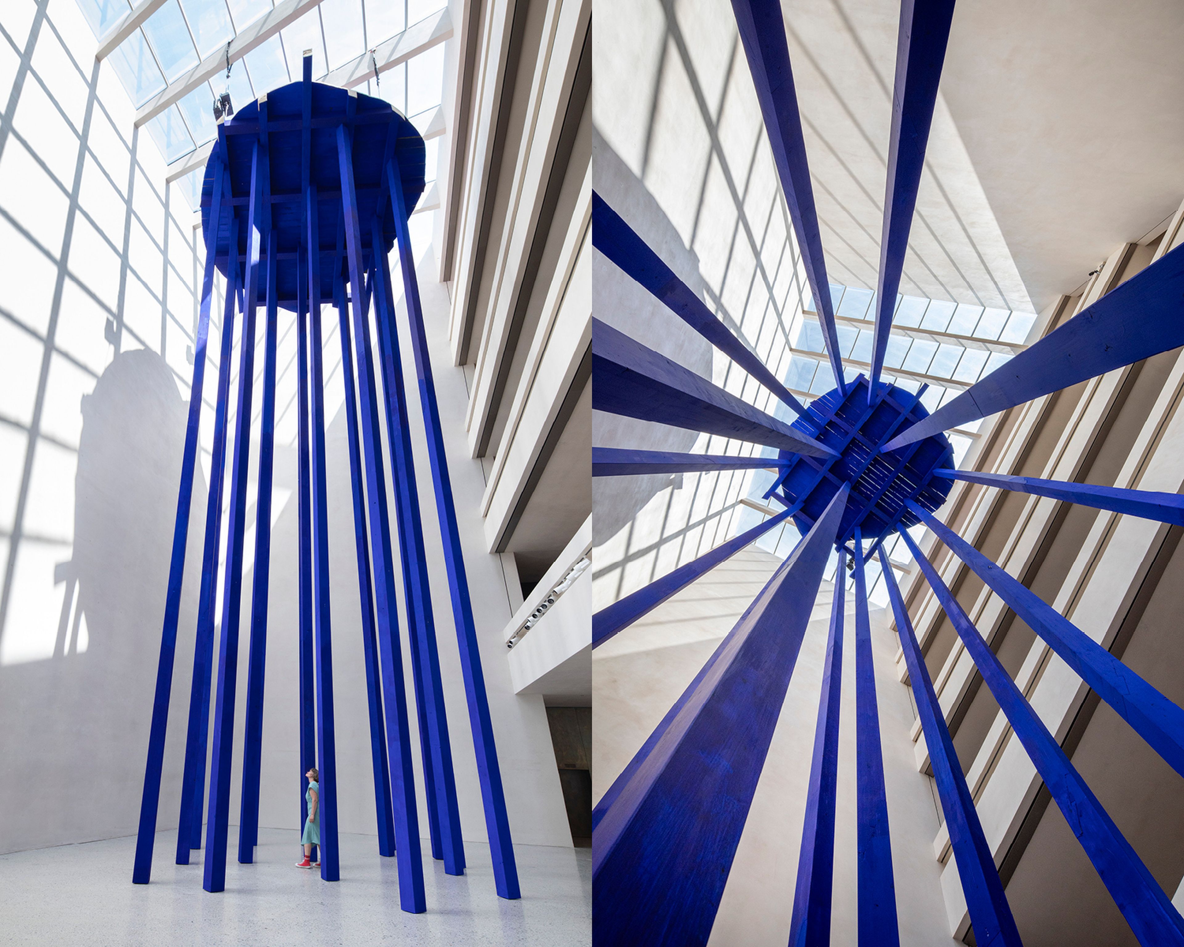 Hoher, blauer Zylinderbau mit vertikalen Trägern in einer hellen Galerie, von unten und von der Seite betrachtet. Oberlichter erzeugen kontrastierende Schatten.