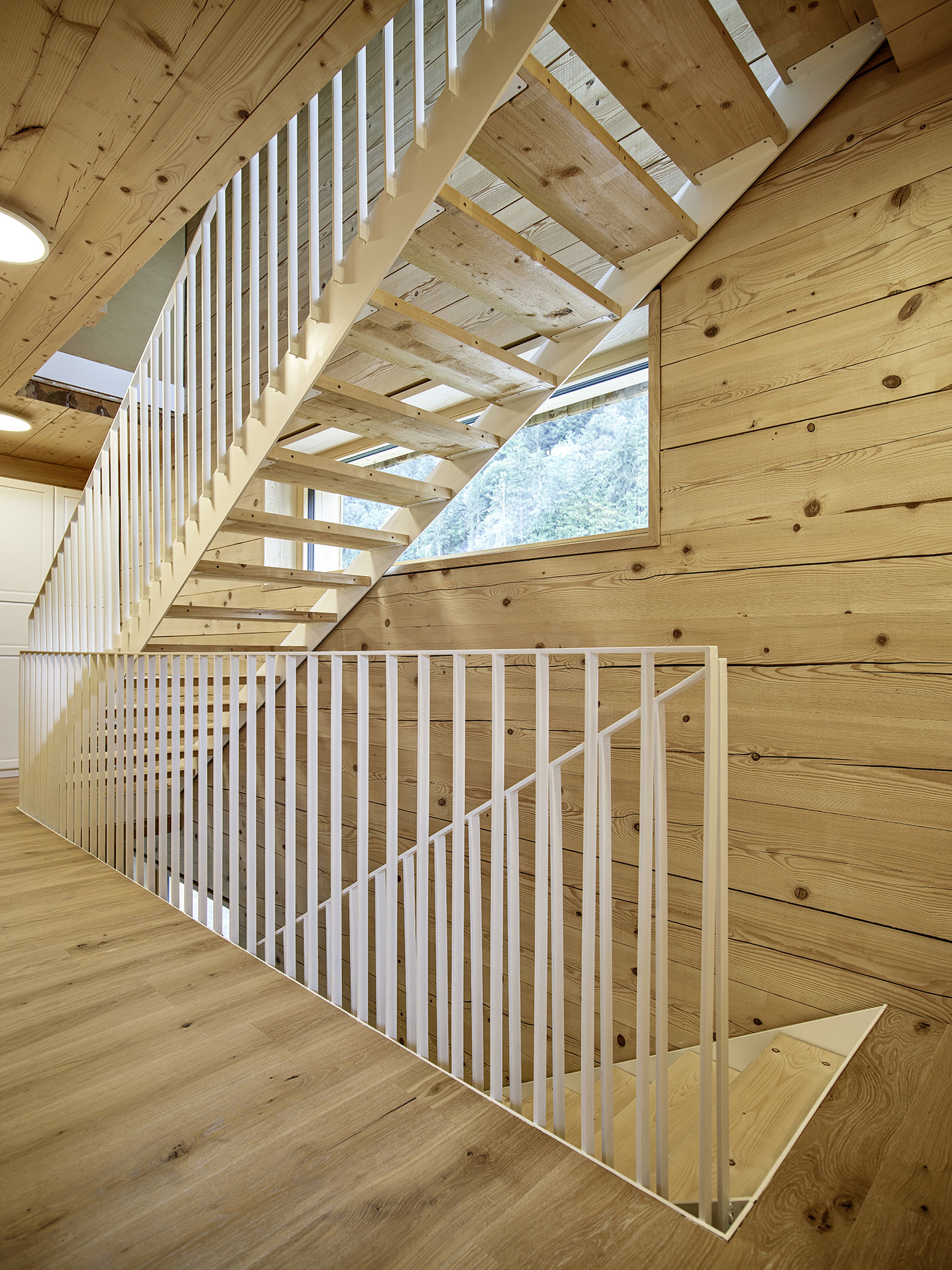 Eine moderne hölzerne Innentreppe mit weißen vertikalen Geländern, angrenzend an ein Fenster mit Blick auf den Wald, die eine natürliche Holzvertäfelung zur Schau stellt.