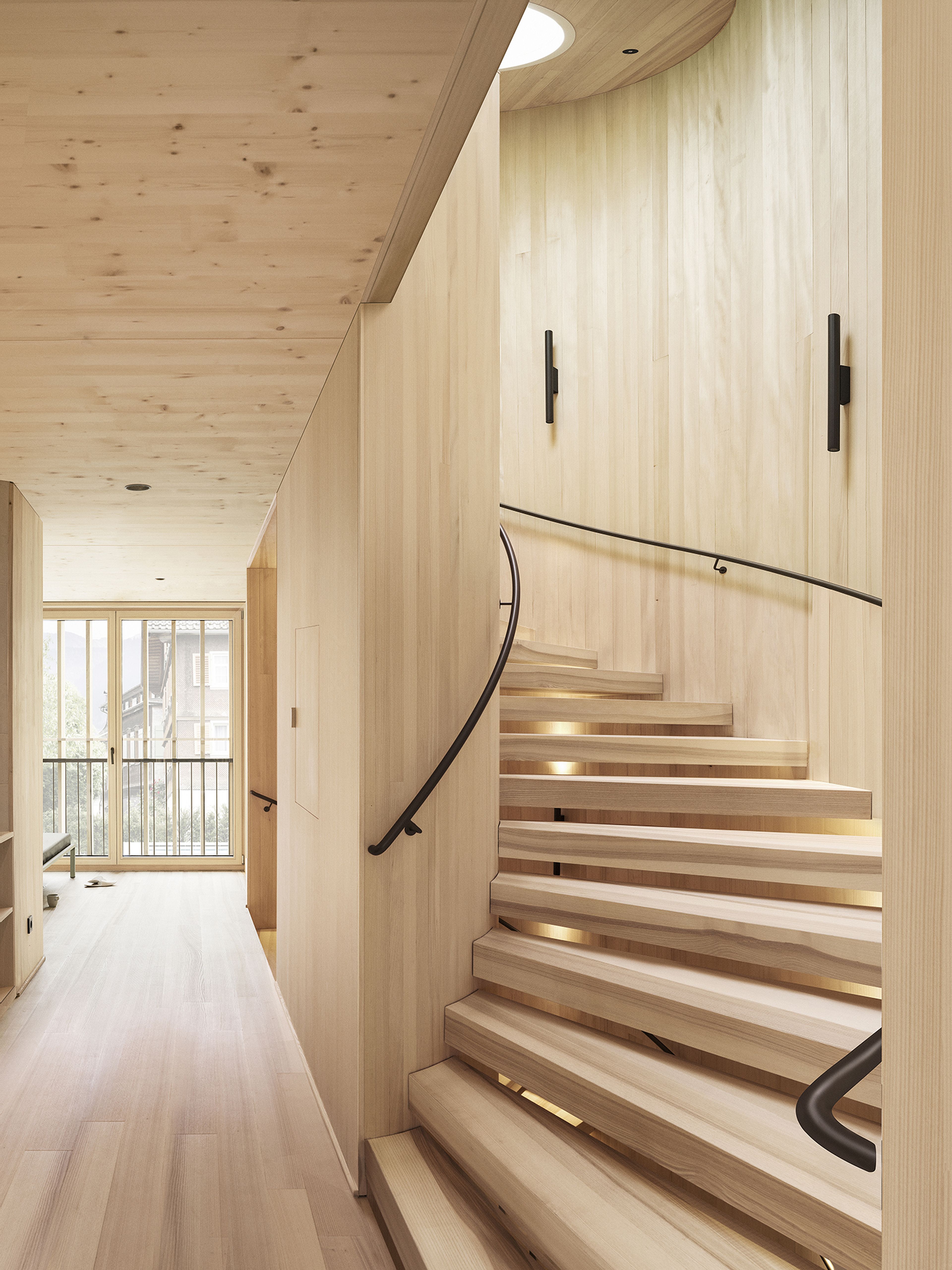 Modernes Interieur aus Holz mit einer Wendeltreppe mit eleganten schwarzen Handläufen und großen Fenstern im Hintergrund, die für natürliches Licht sorgen.