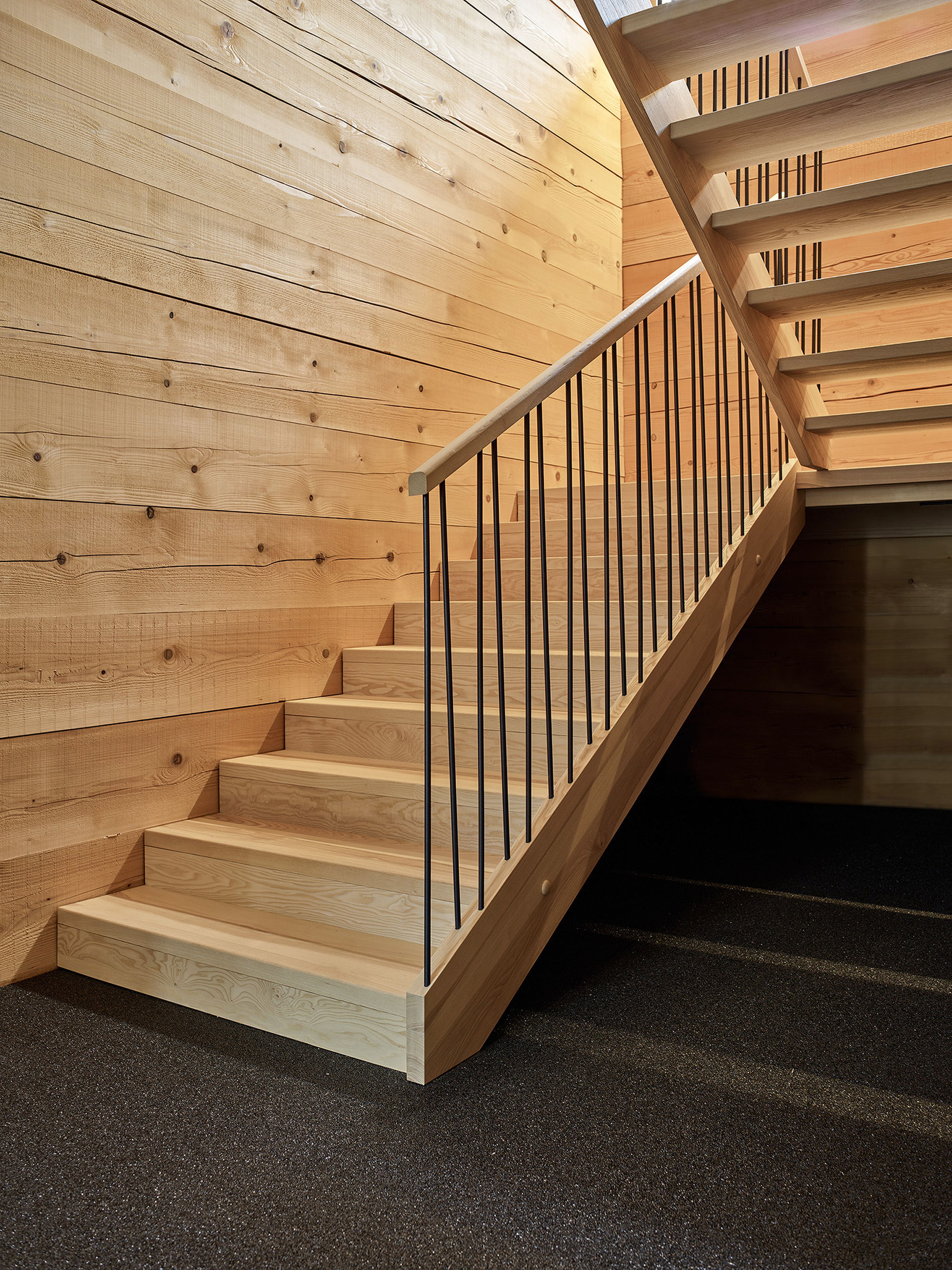 Eine Holztreppe mit Metallgeländer steht vor einer Wandverkleidung aus Holzpaneelen. Die Treppe führt nach oben, wo sich ein teilweise sichtbarer Treppenabsatz befindet.