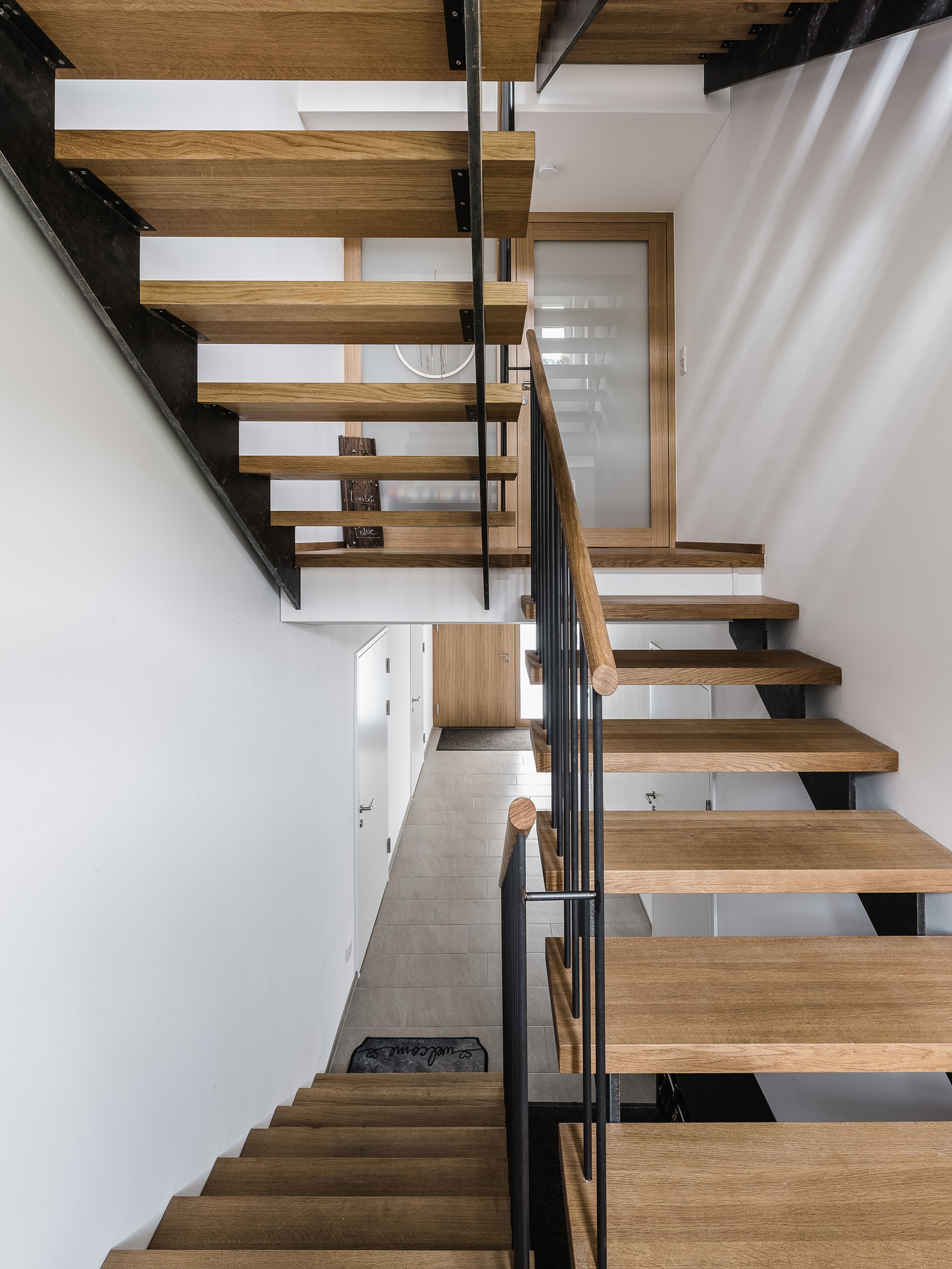 Modernes Interieur mit einer Treppe aus Holz und Metall, die sich durch minimalistisches Design und elegante Geländer auszeichnet und zu einem weißen, hellen Flur führt.