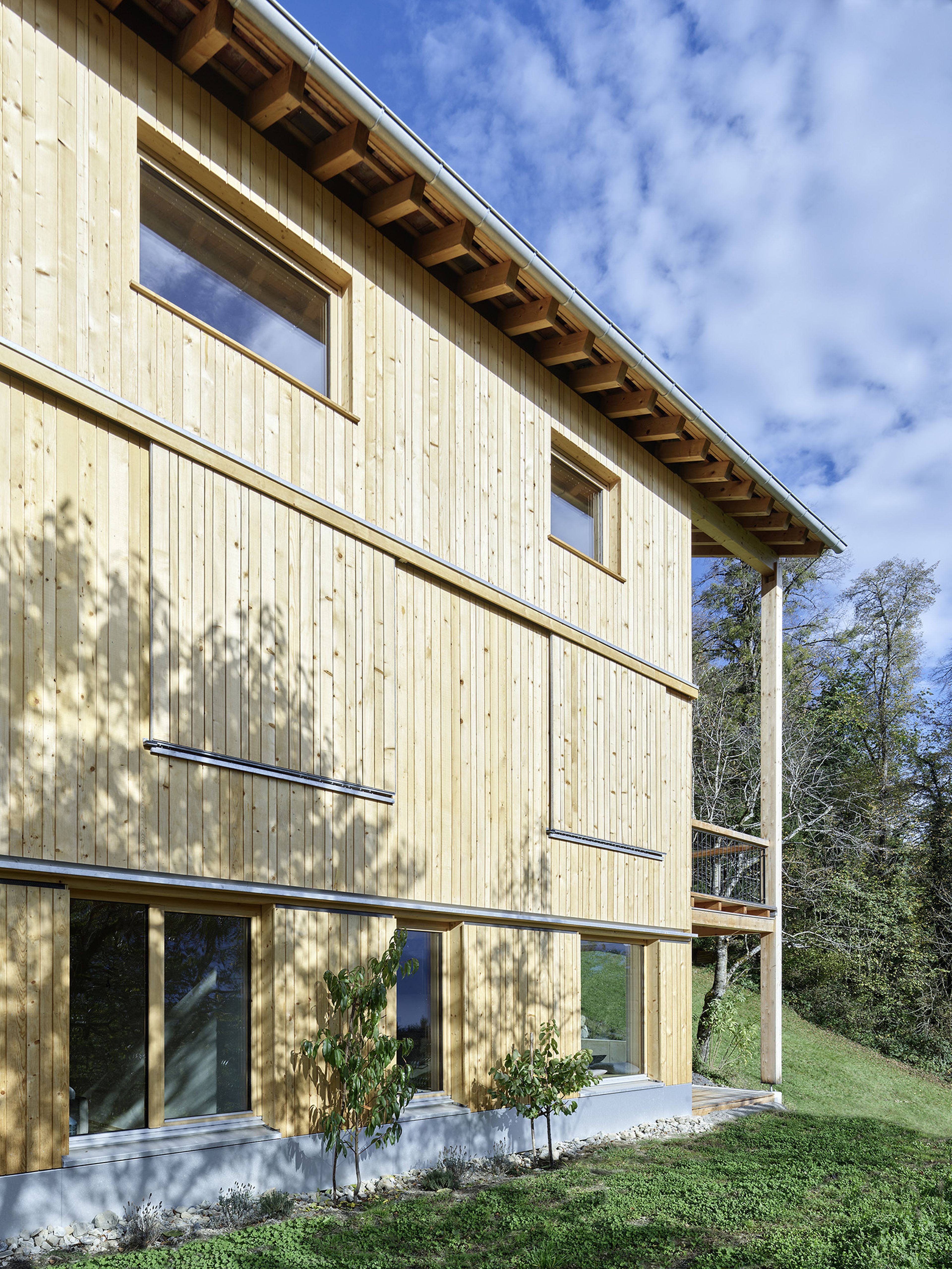 Ein Holzgebäude mit großen Fenstern und weit überhängendem Dach, vor blauem Himmel und Bäumen. Schatten und Sonnenlicht betonen die natürliche Struktur.