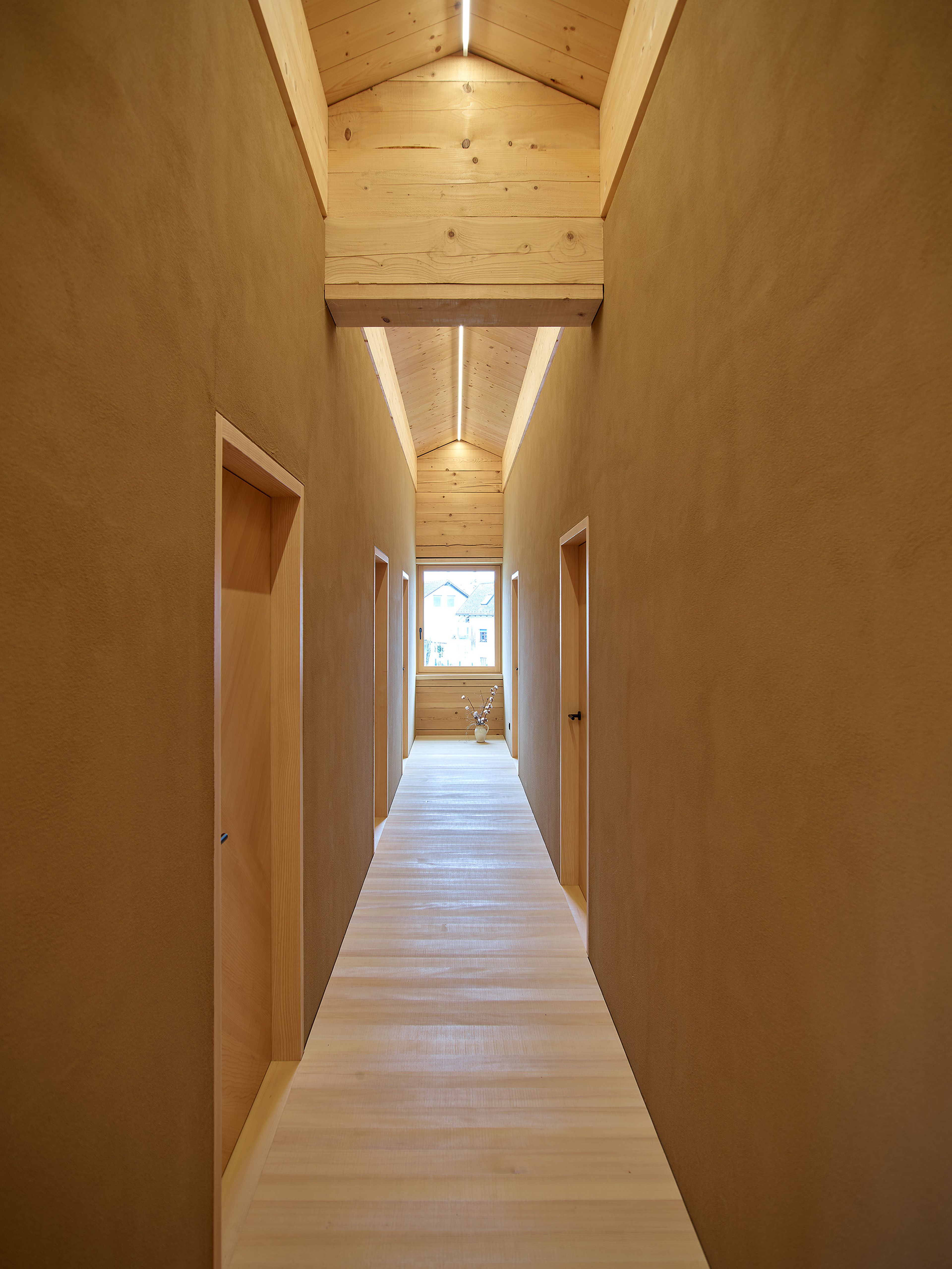 Foto eines Gangs in einem Einfamilienhaus in gestrickter, massiver Holzbauweise mit sichtbaren Holzbalken, Lehmputzwänden, warmem Naturlicht und natürlicher, ruhiger Wohnatmosphäre.
