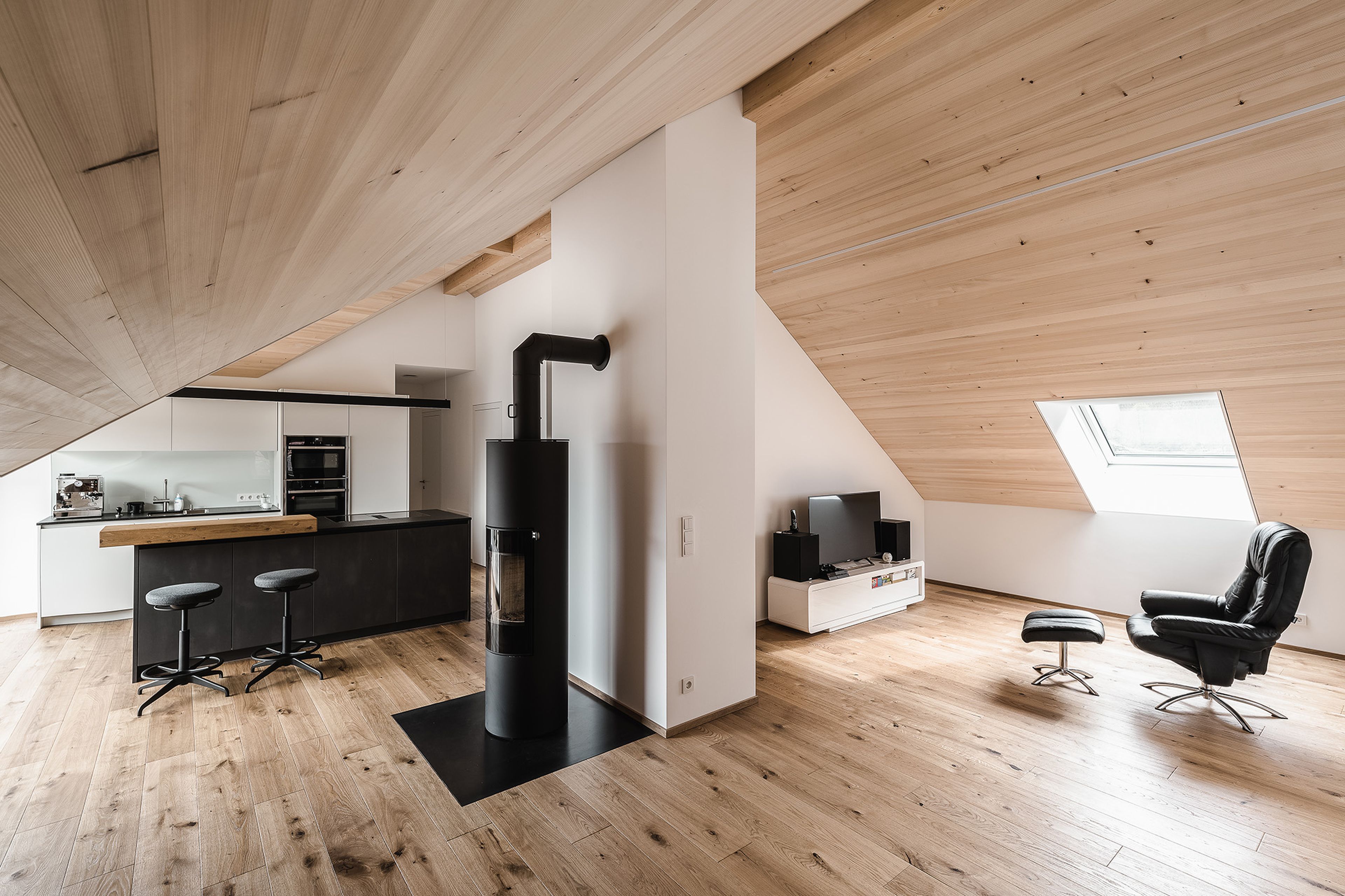 Innenansicht eines renovierten Dachausbaus in Holz mit Holzdecke und Parkettboden und moderner Küche in Schwarz.
