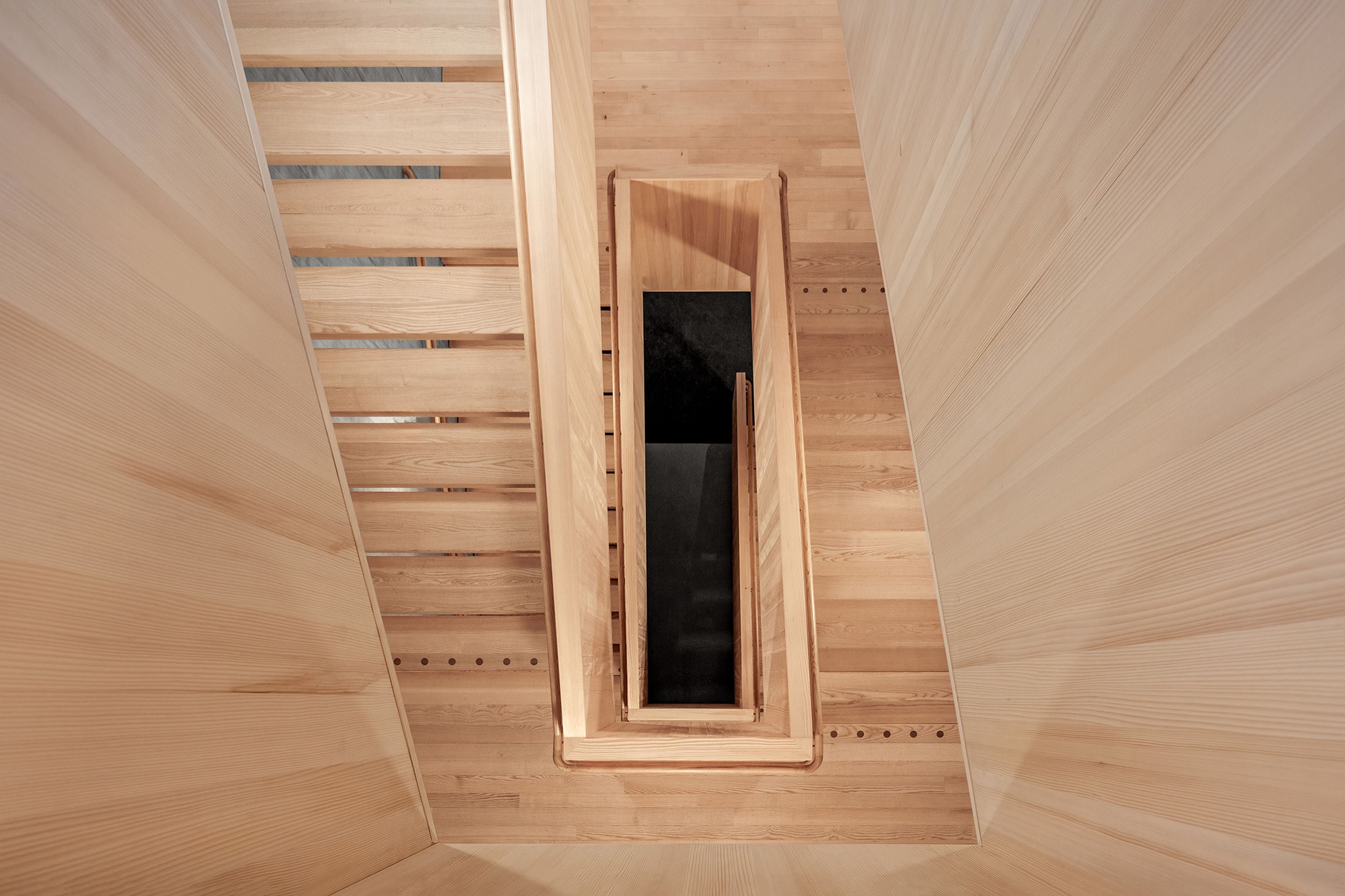 Eine hölzerne Treppe mit parallelen Treppenläufen vor einer grauen Betonwand, die vom Sonnenlicht durch ein großes Fenster erhellt wird.