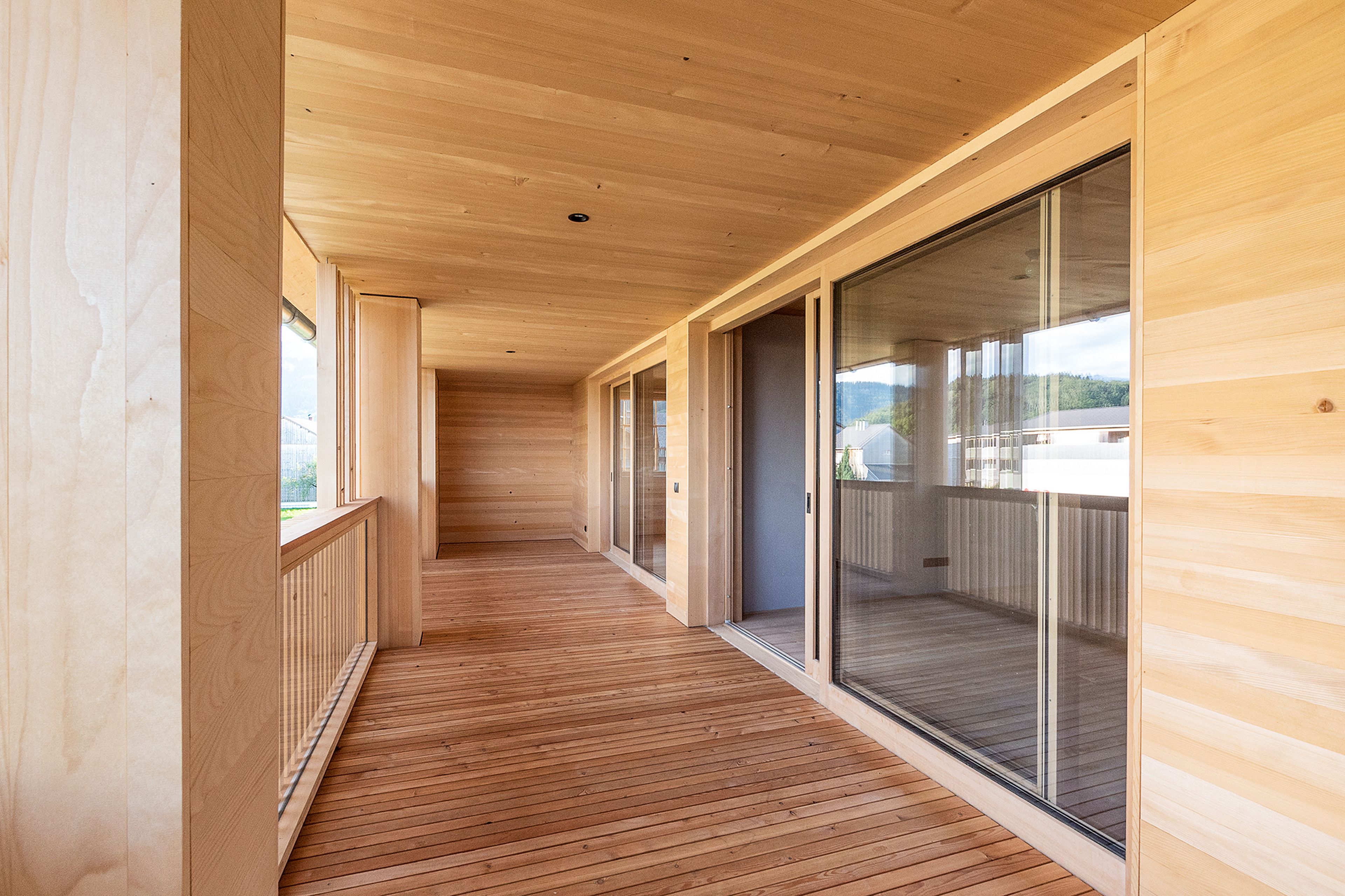 Helle Holzterrasse mit großen Glasschiebetüren auf der rechten Seite und einem Holzgeländer auf der linken Seite, der von natürlichem Licht erhellt wird.