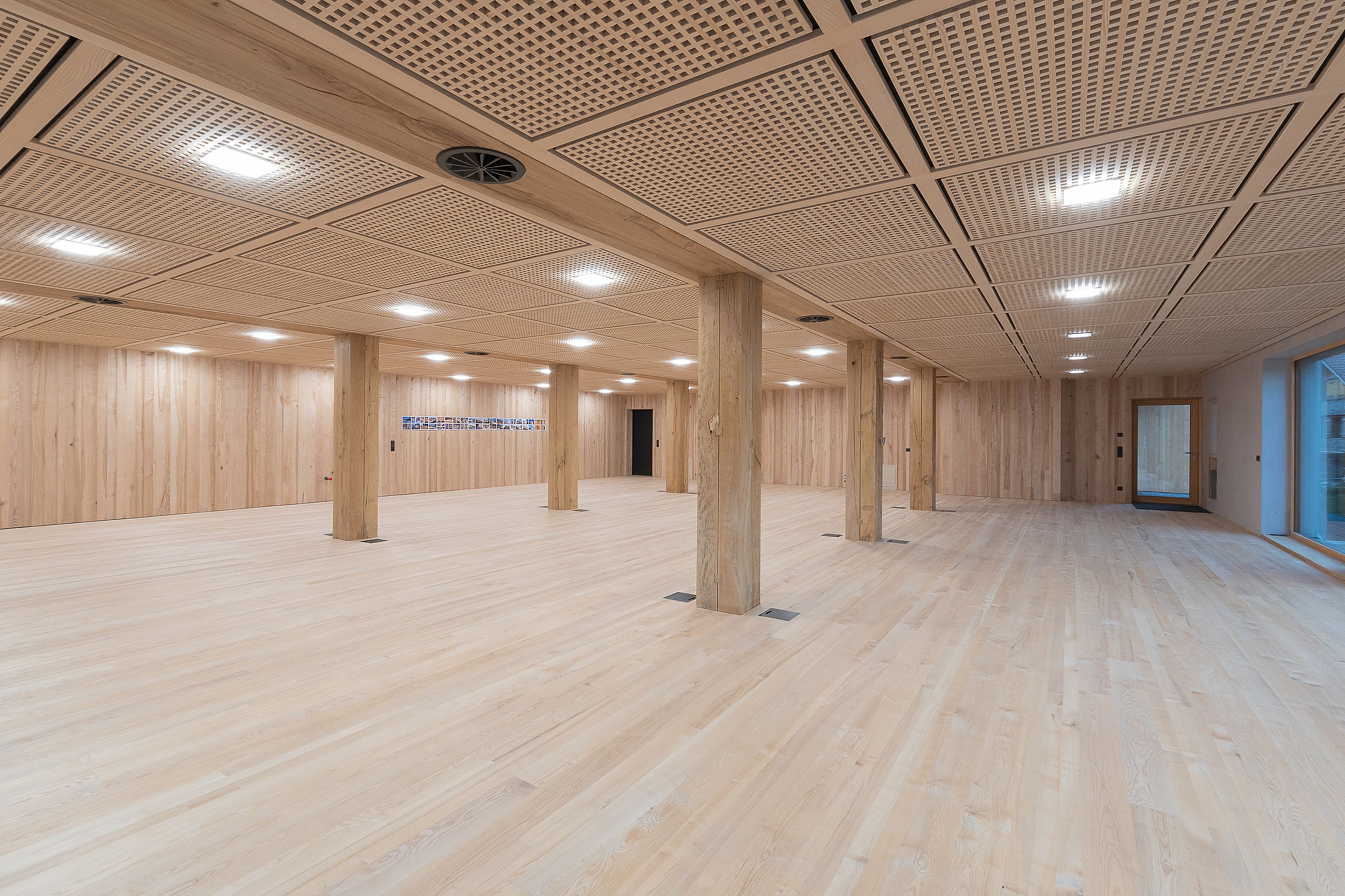 Großzügiger Raum mit Holzboden und -wänden, Deckenleuchten und mehreren hölzernen Stützsäulen. Große Fenster auf der rechten Seite.