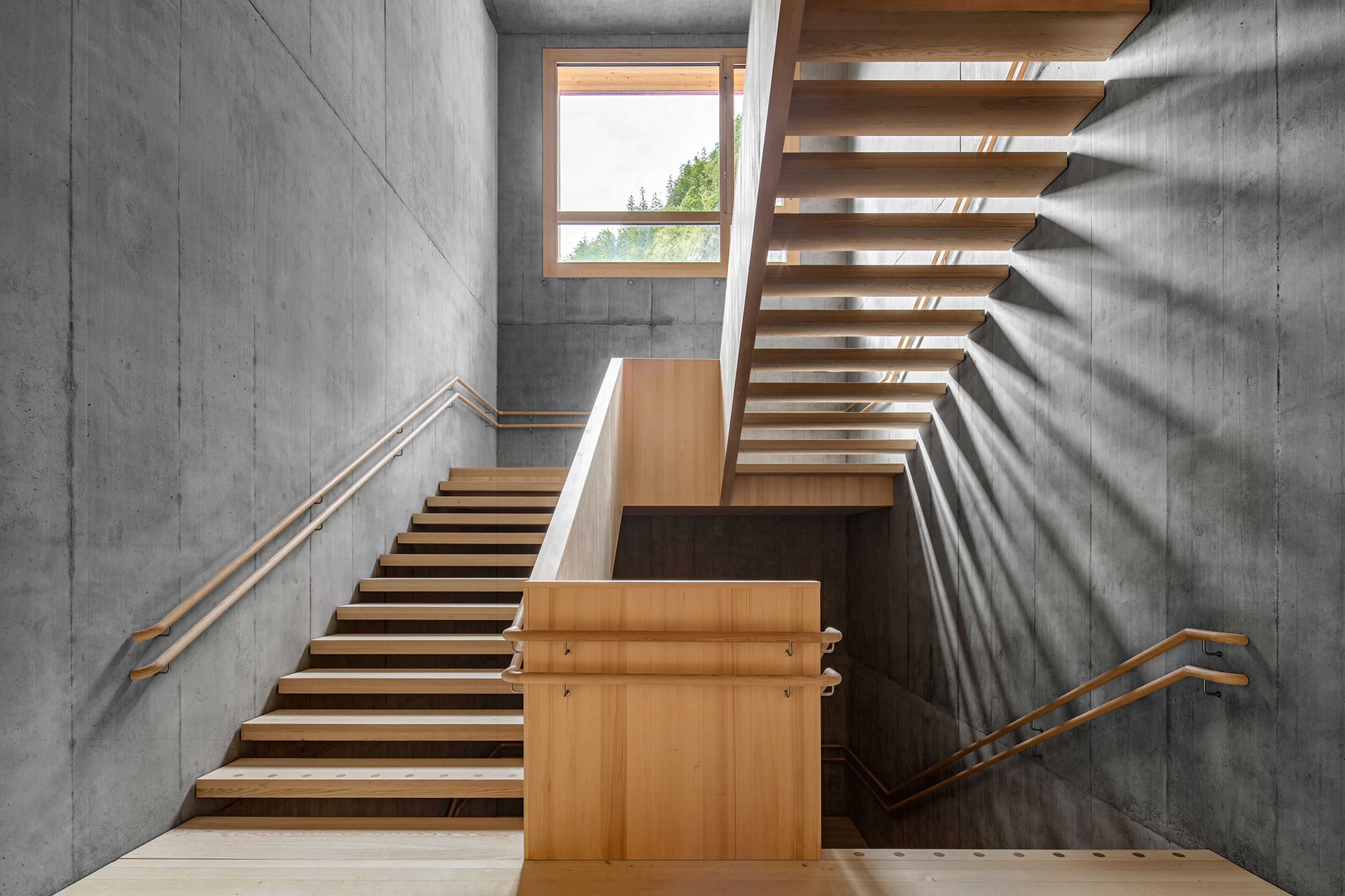 Eine hölzerne Treppe mit parallelen Treppenläufen vor einer grauen Betonwand, die vom Sonnenlicht durch ein großes Fenster erhellt wird.