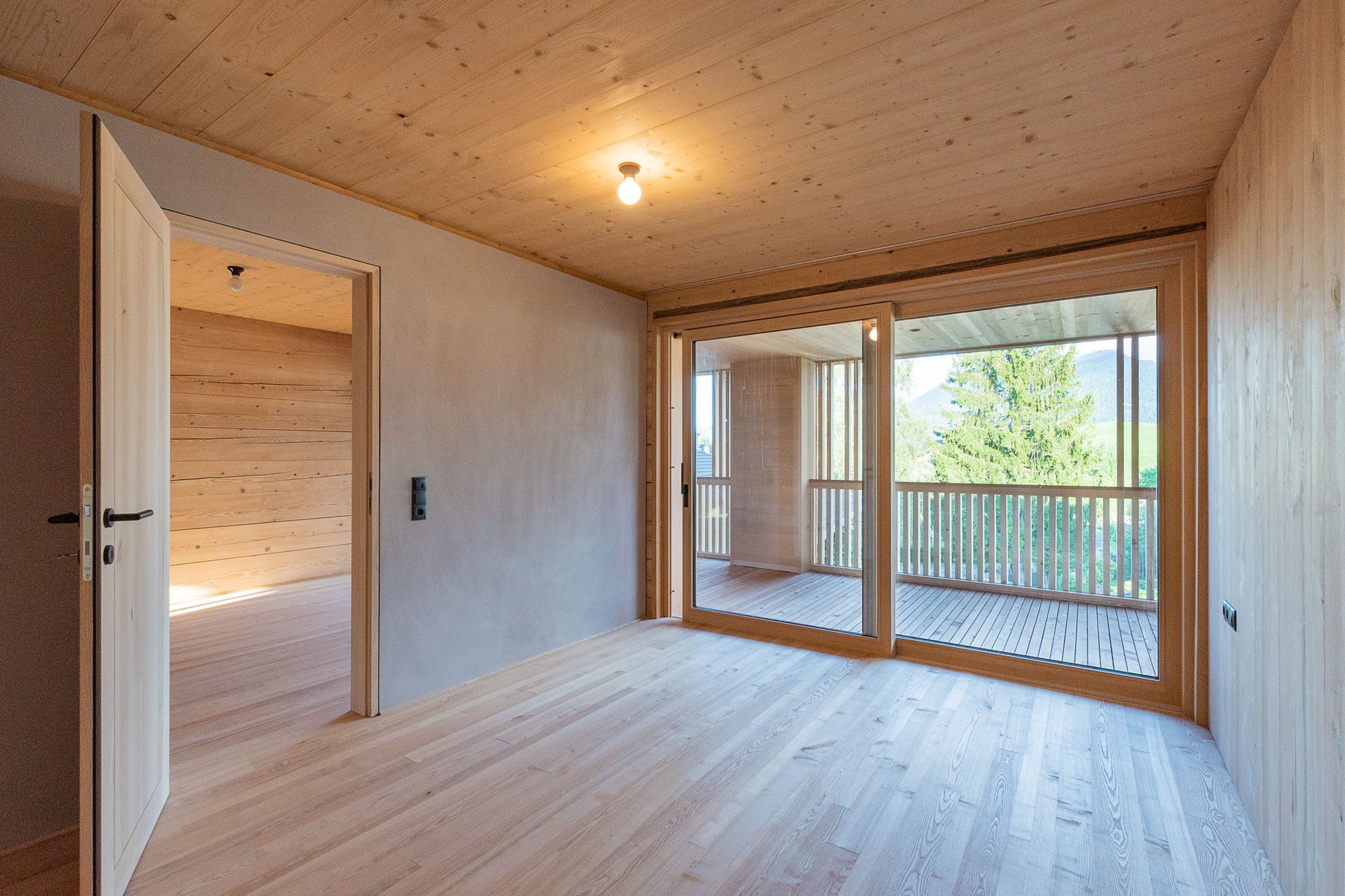 Ein helles, leeres Zimmer mit Holzboden und Holzdecke, offener Tür zu einem anderen Zimmer und großen Fenstern, die auf einen Balkon mit Blick auf Bäume führen.