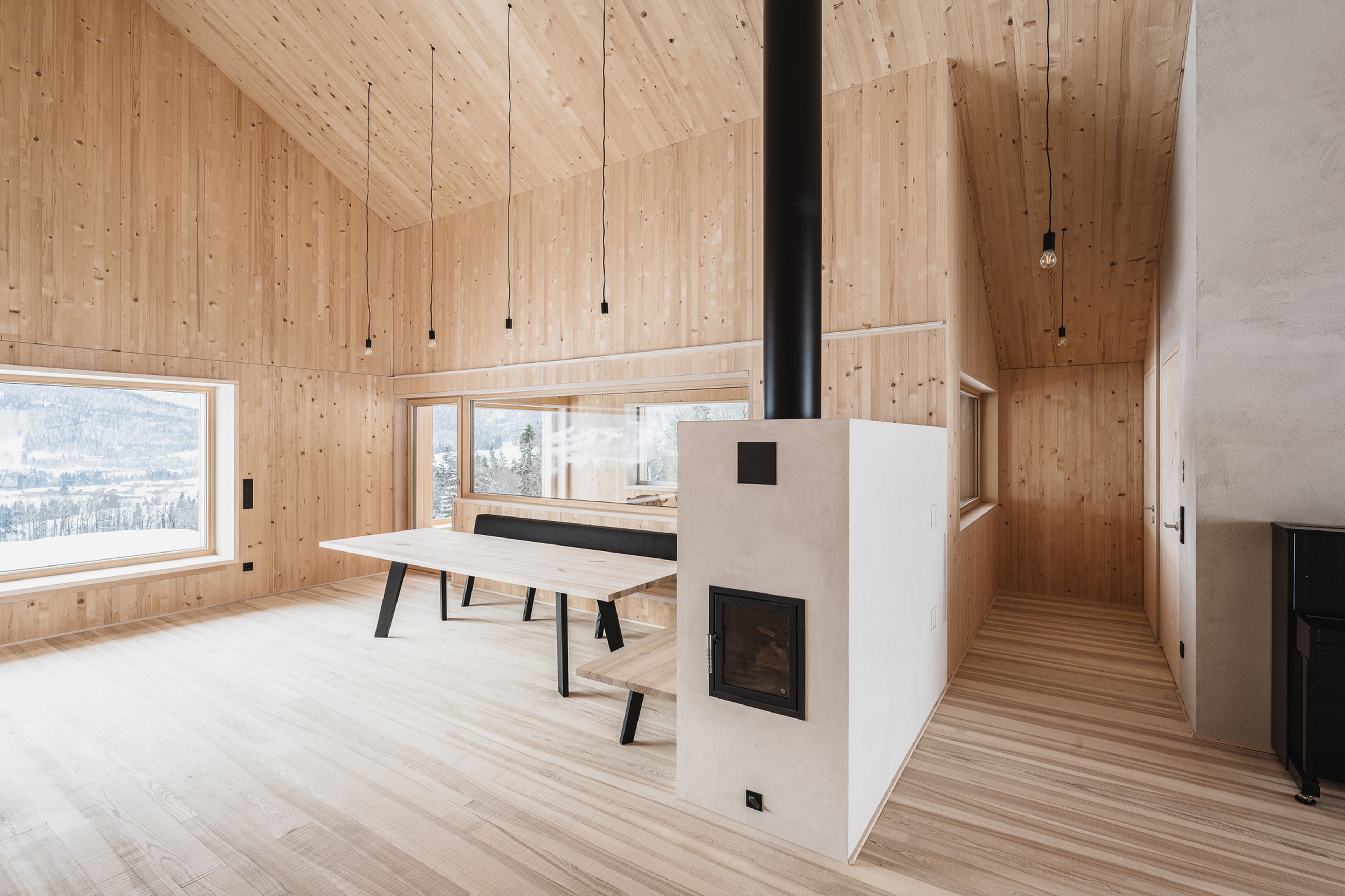 Großzügiger, minimalistischer Holzinterieur mit hoher Decke, großem Fenster, Esstisch und Kamin. Helle Holztöne dominieren den Raum.