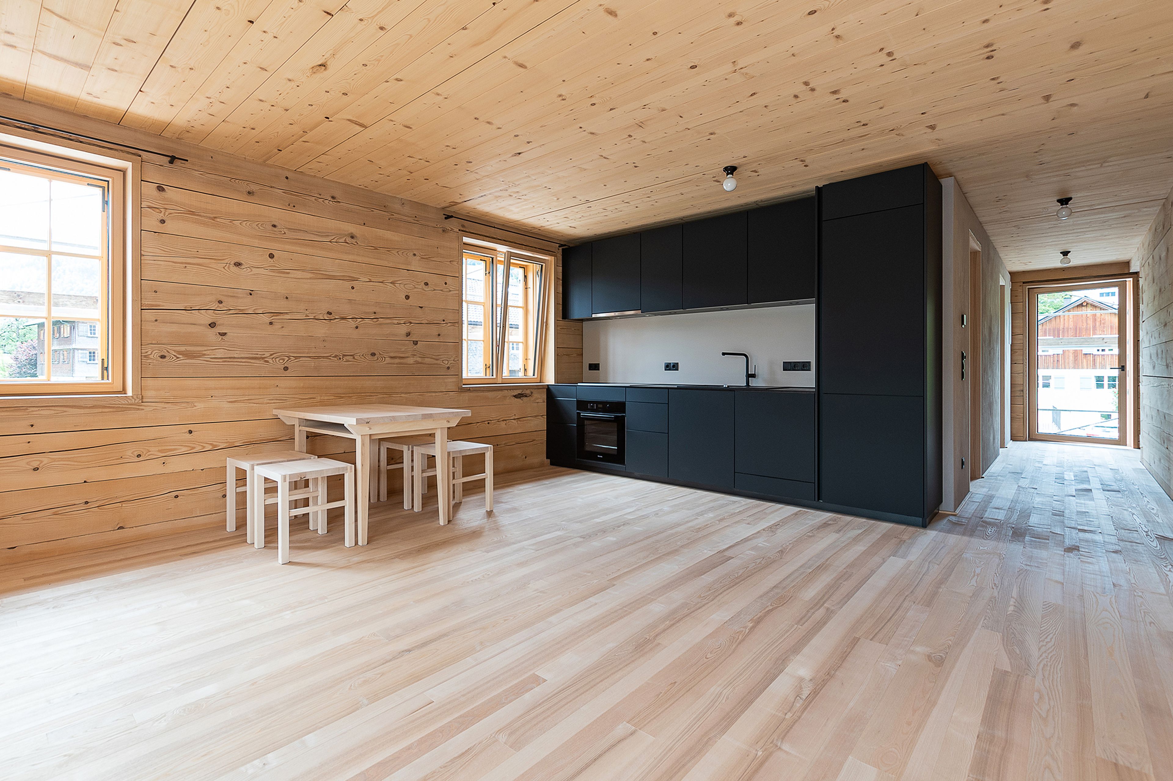 Minimalistisches Interieur aus Holz mit schwarzer Küche, hellen Holzböden und einem kleinen Esstisch in Fensternähe. Heller, offener Raum.
