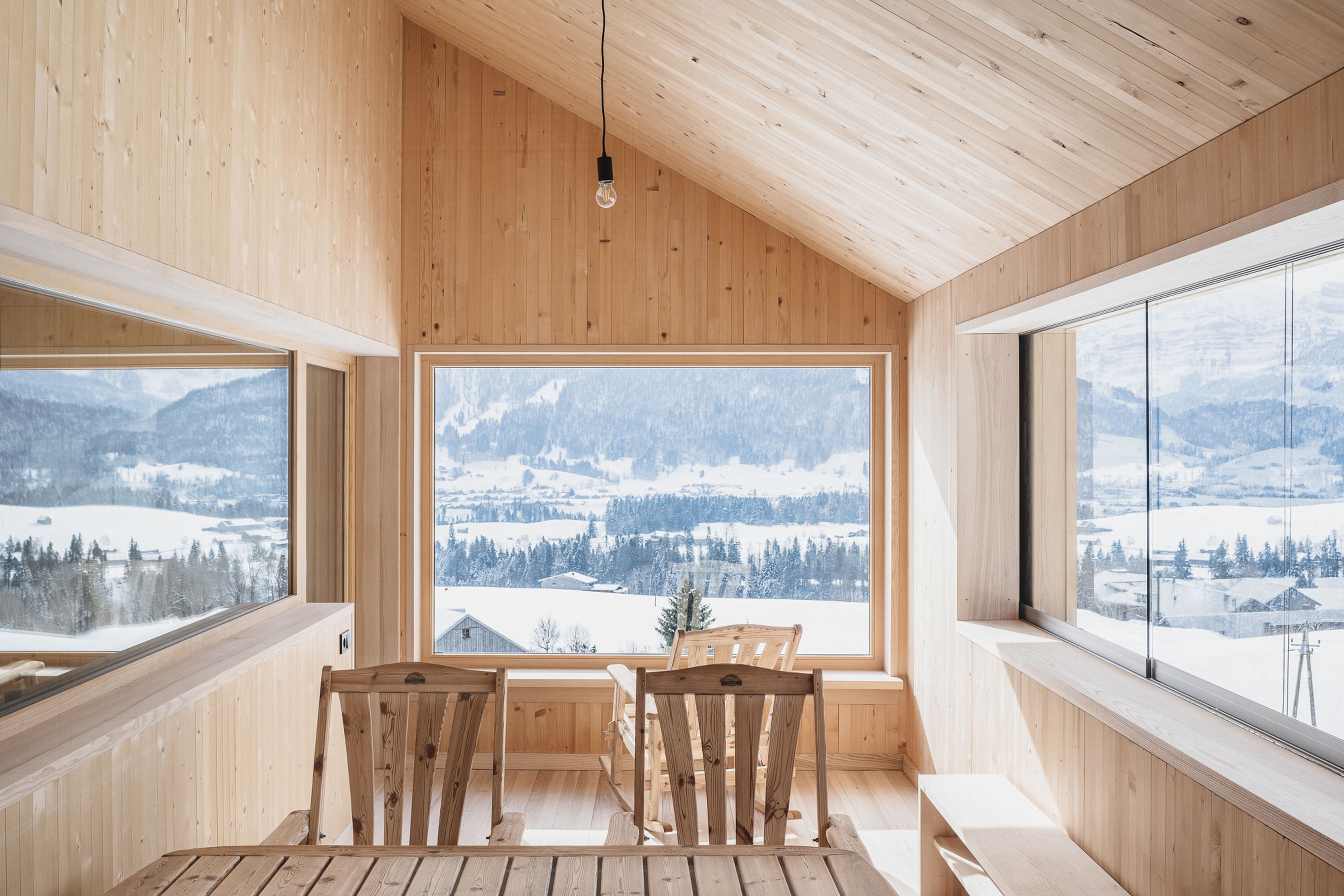 Wood-paneled room with large windows offering snowy mountain views, featuring a simple light bulb, wooden chairs, and a spacious table.