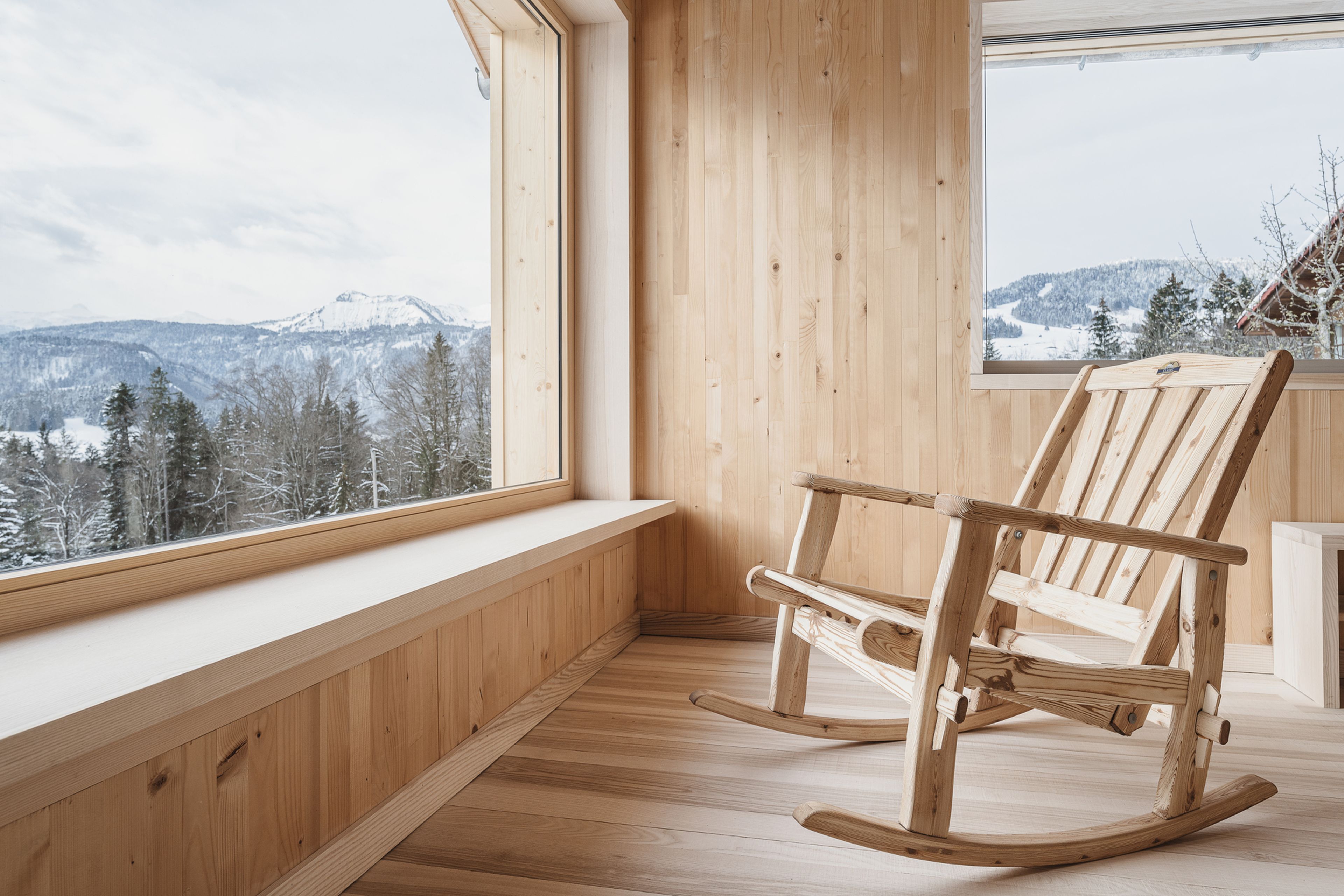 Wooden room with a rocking chair by large windows, overlooking snowy mountains and pine trees. Natural light brightens the cozy space.