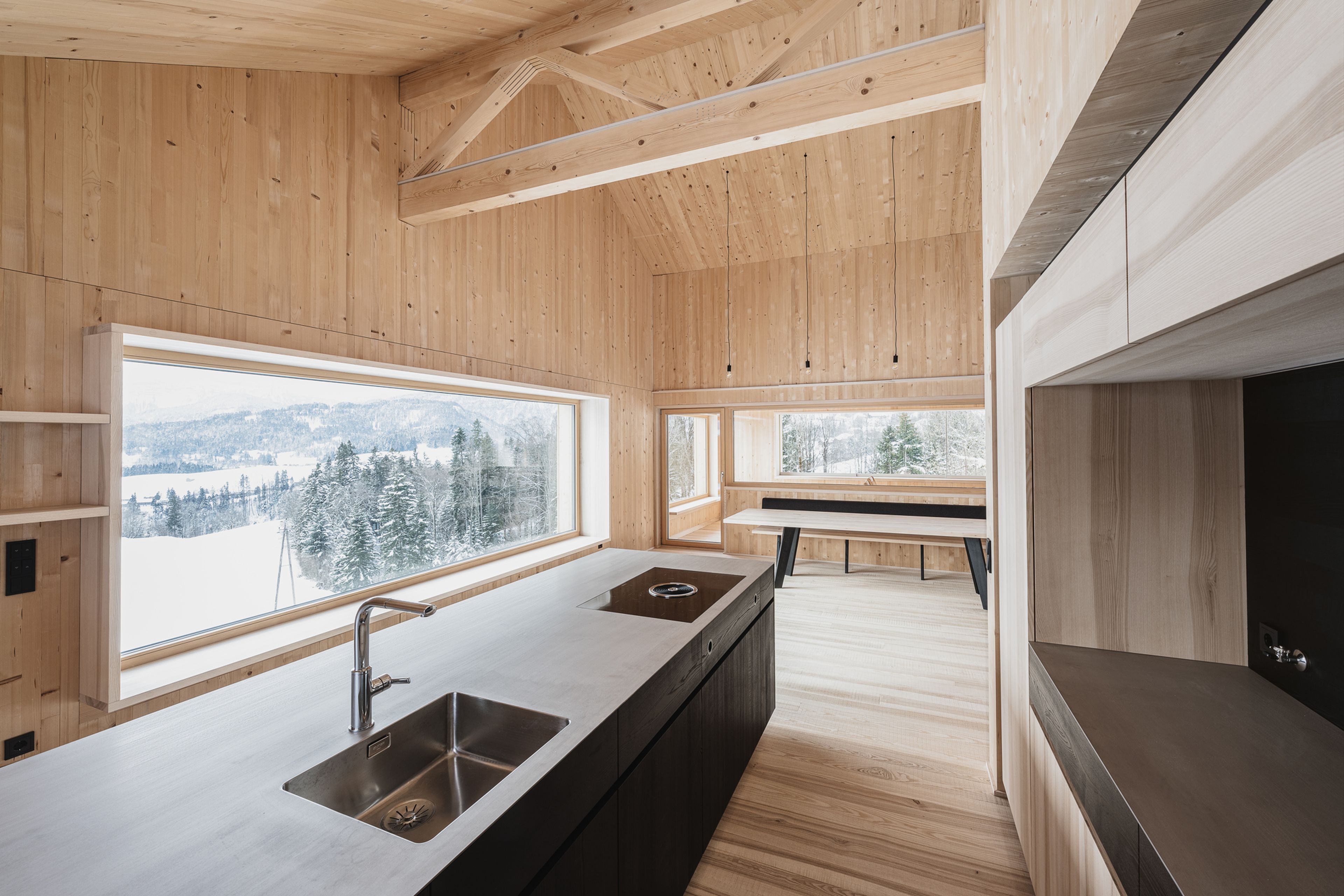 Minimalistische Küche und Essbereich mit hellen Holzvertäfelungen und großen Fenstern mit Blick auf den verschneiten Wald. Schwarze Arbeitsplatten runden das Gesamtbild ab.