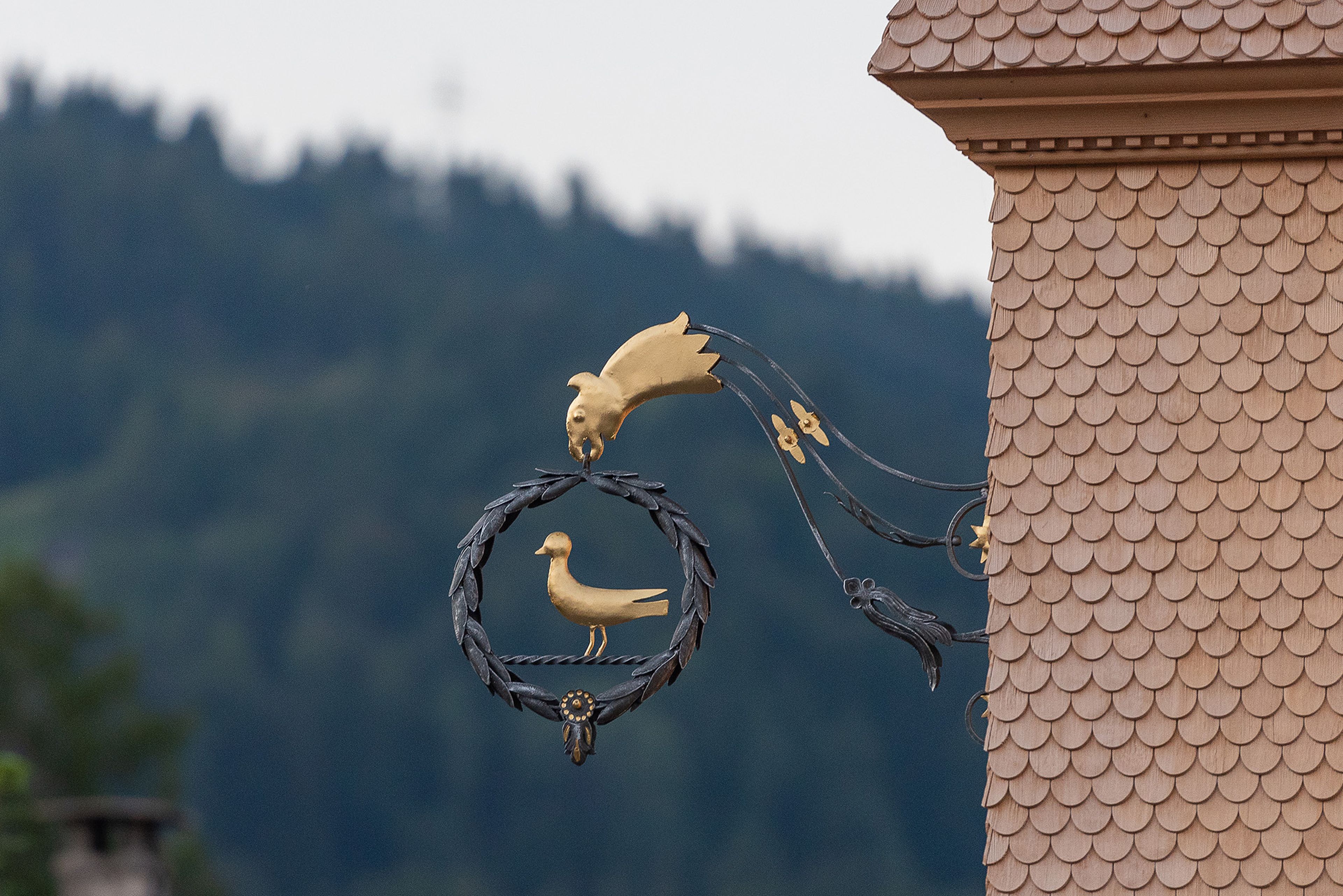 Ornate metal decoration on building facade with gold bird and leaf design, set against a blurred mountainous background.