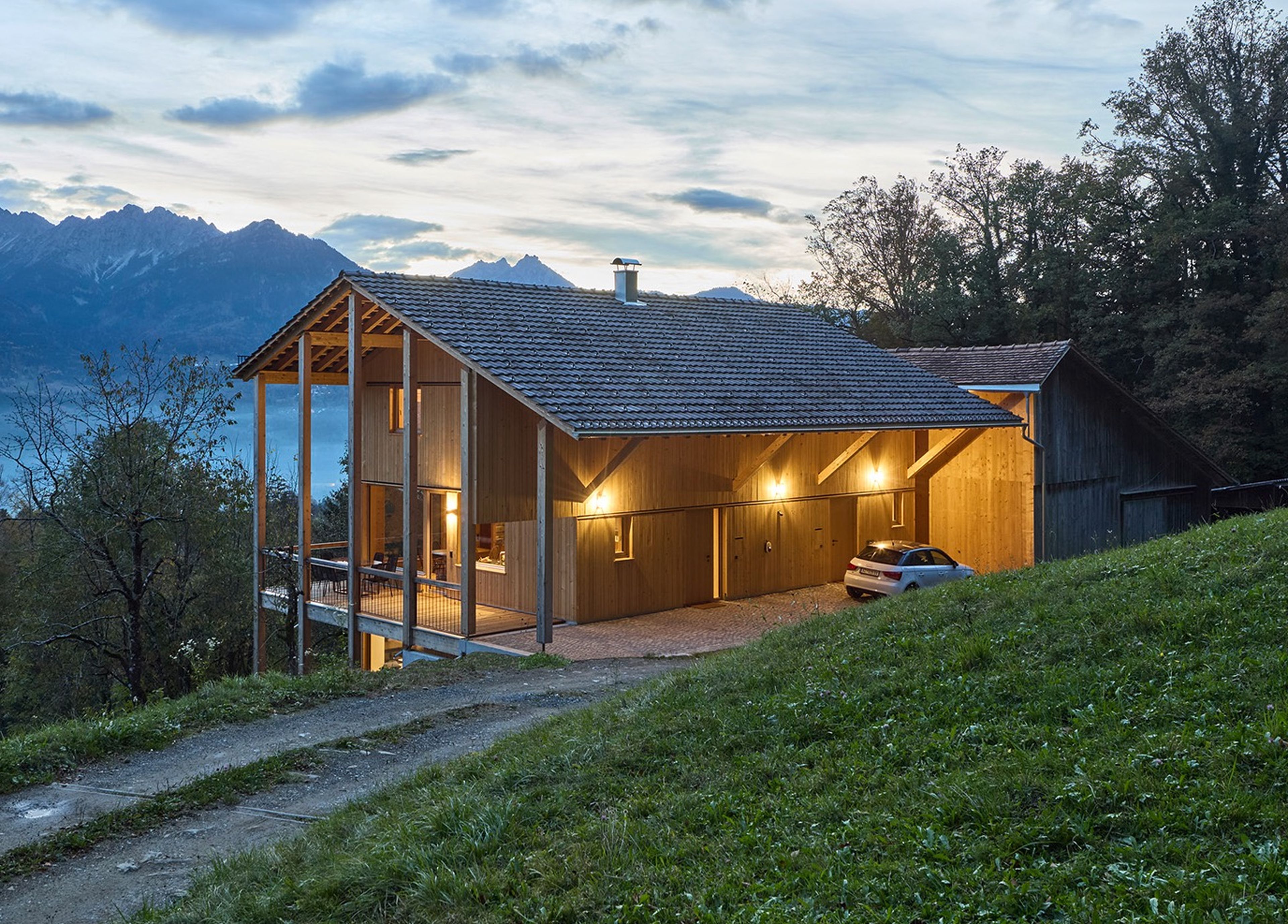 Modernes Holzhaus mit beleuchteter Veranda, gelegen auf einem grasbewachsenen Hügel mit Blick auf ein Tal und die Berge in der Abenddämmerung. Auto neben dem Haus geparkt.