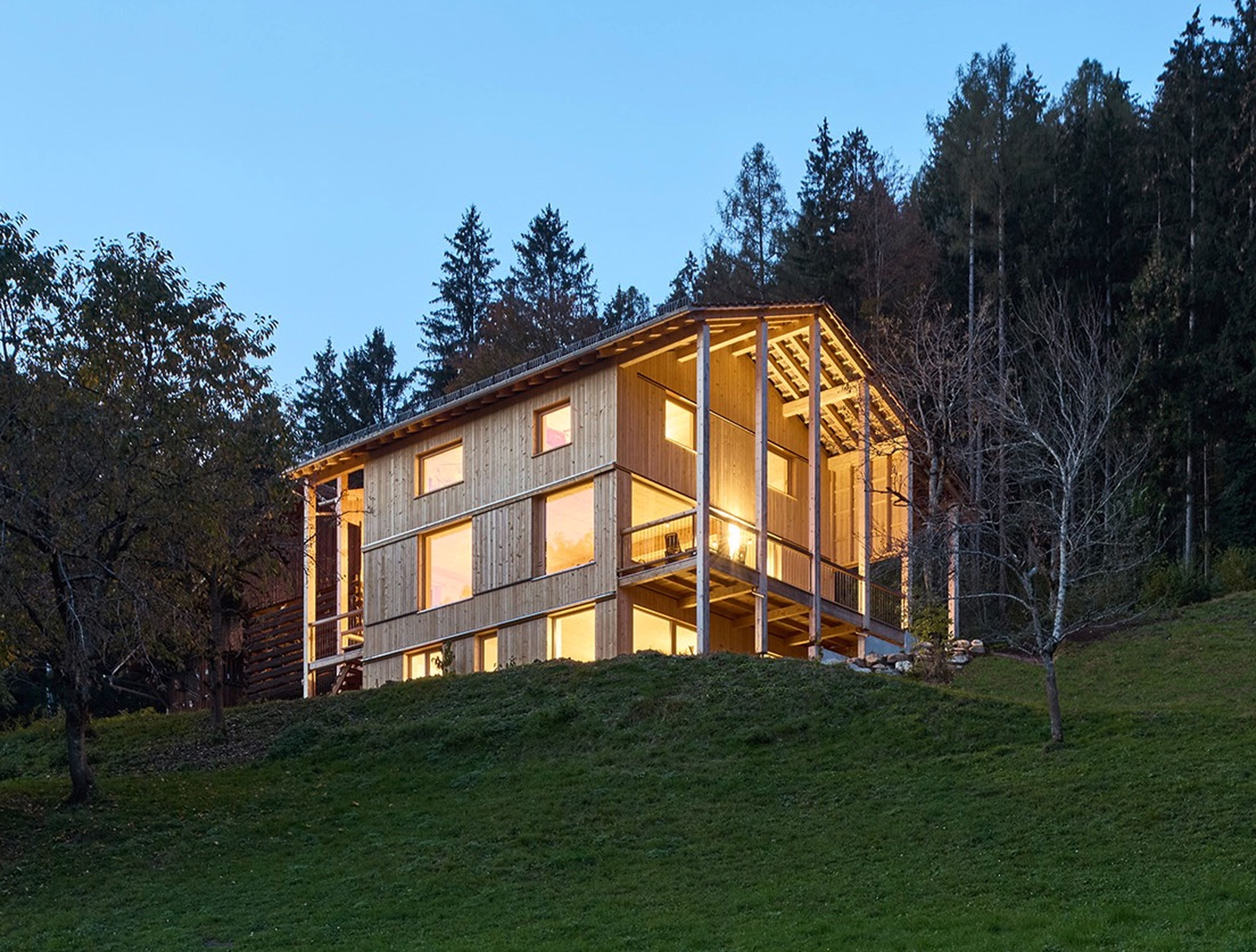 Ein modernes Holzhaus mit großen Fenstern wird in der Abenddämmerung beleuchtet, umgeben von grasbewachsenen Hängen und dichtem Baumbestand.