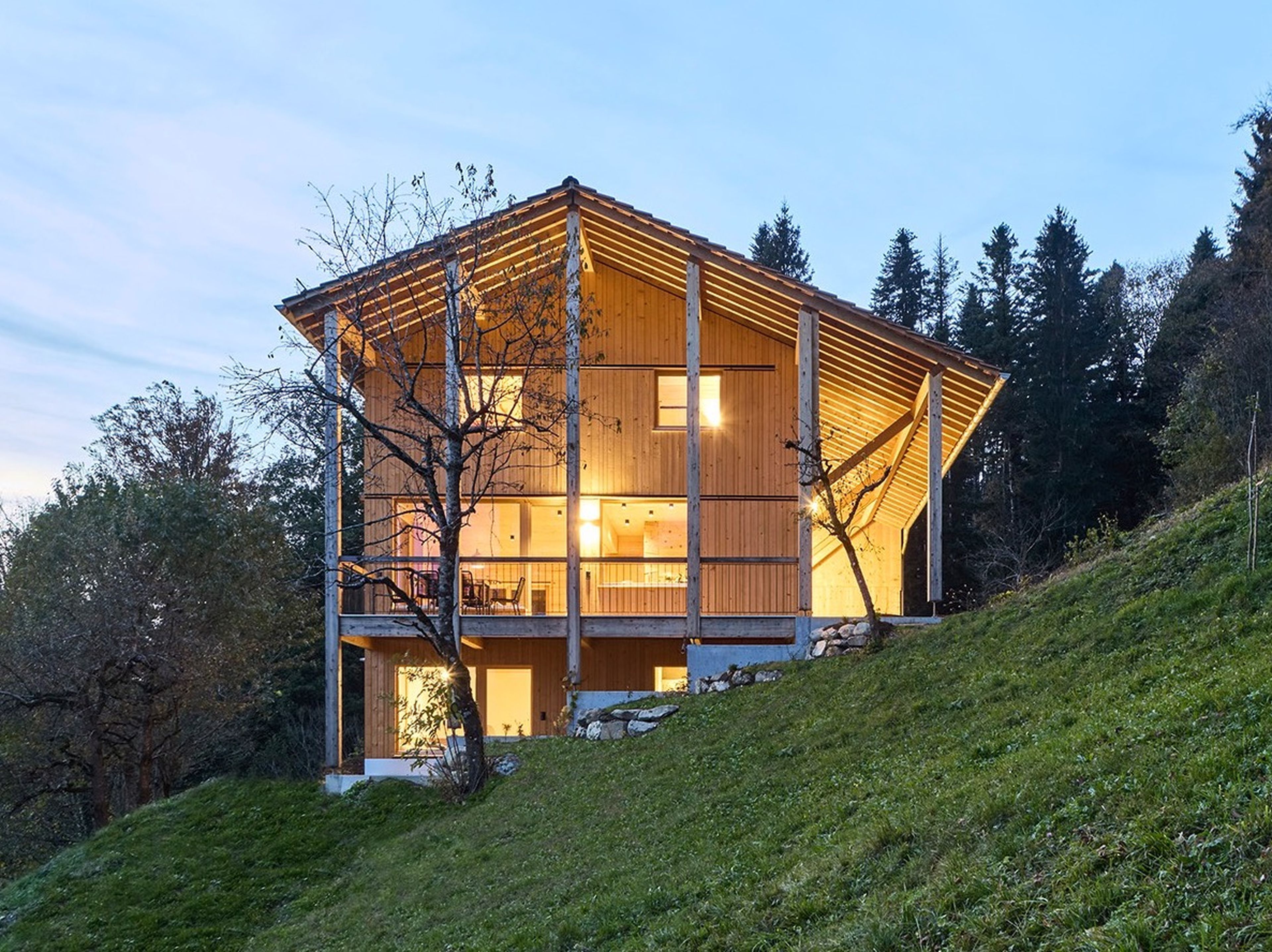 Ein modernes Holzhaus mit großen Fenstern an einem grasbewachsenen Hang, umgeben von Bäumen in der Abenddämmerung, mit warm leuchtenden Innenlichtern.