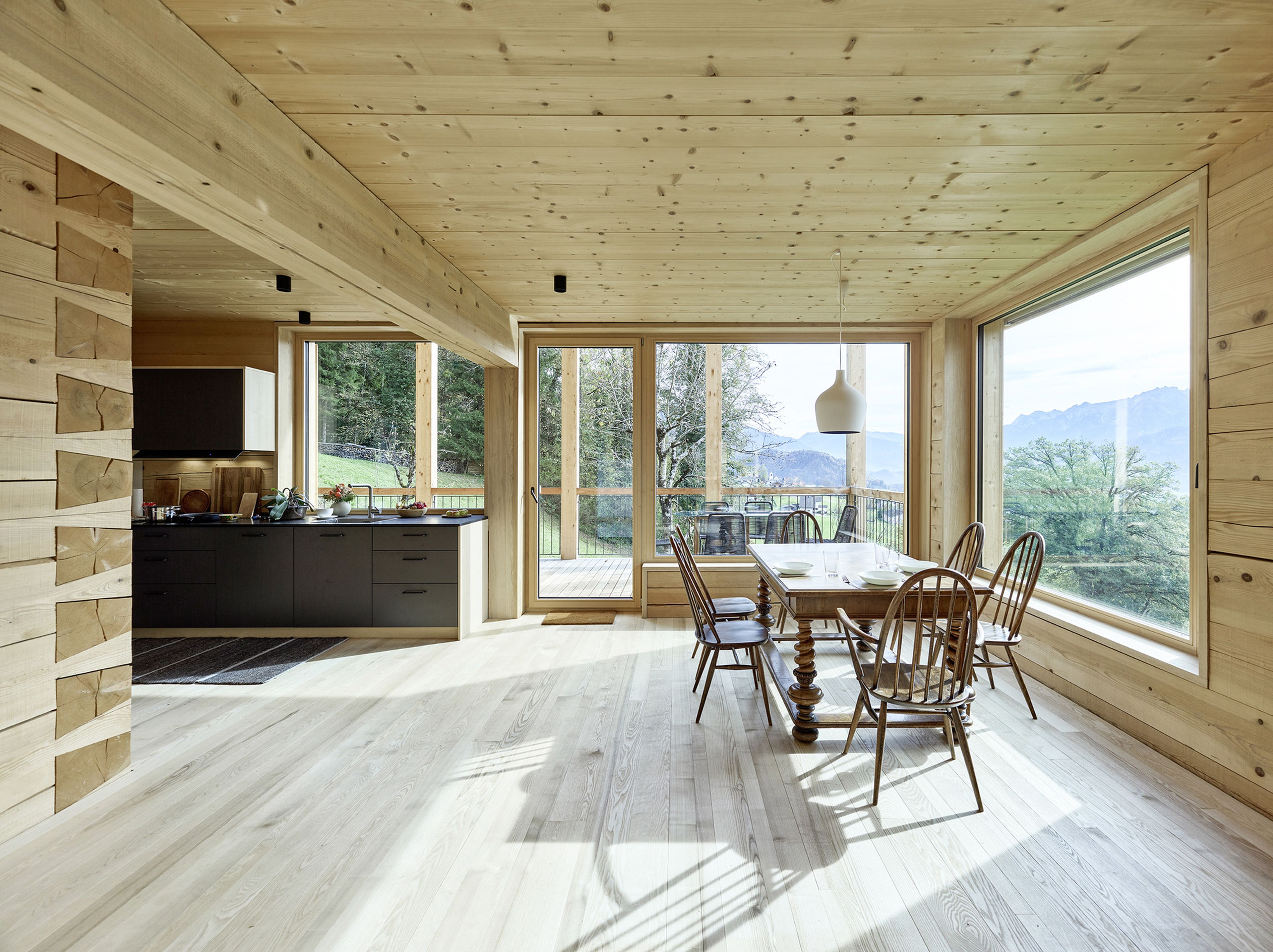 Essbereich mit Holzverkleidung, großen Fenstern mit Bergblick, einem Holztisch mit Stühlen und einer Küche mit dunklen Schränken.