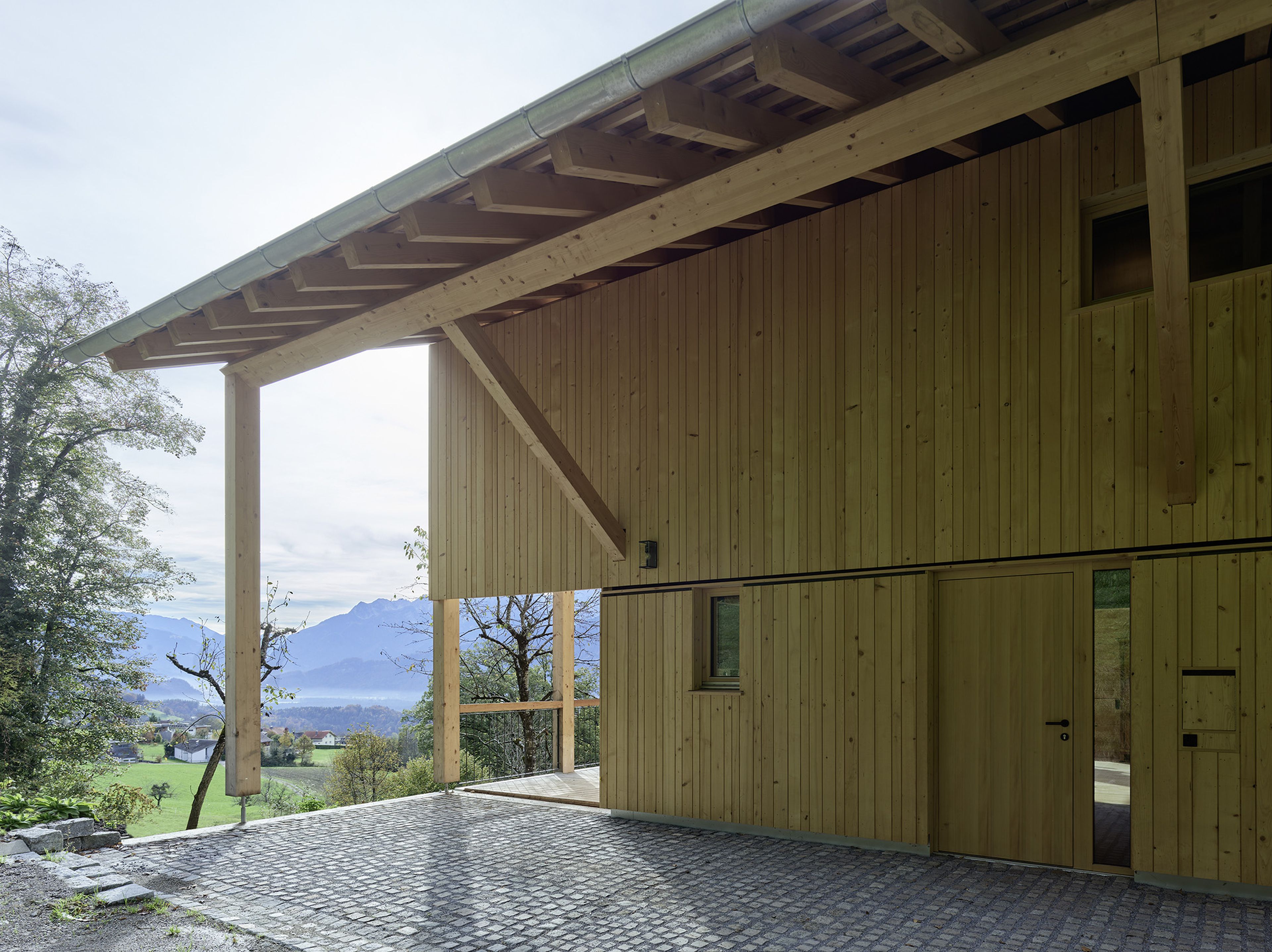 Holzhaus mit großem Vordach, das einen malerischen Blick auf ein Tal mit Bergen und Bäumen bietet. Im Vordergrund ein Kopfsteinpflasterweg.